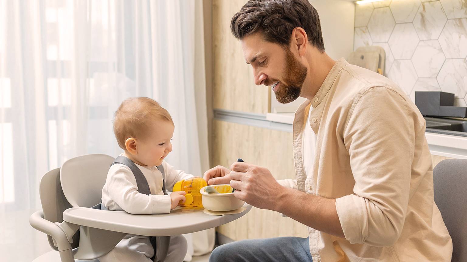 Vater füttert Baby zu Hause im Hochstuhl. Das Baby hält ein Spielzeug, während der Vater ihm einen Löffel Essen aus einer Schüssel anbietet.