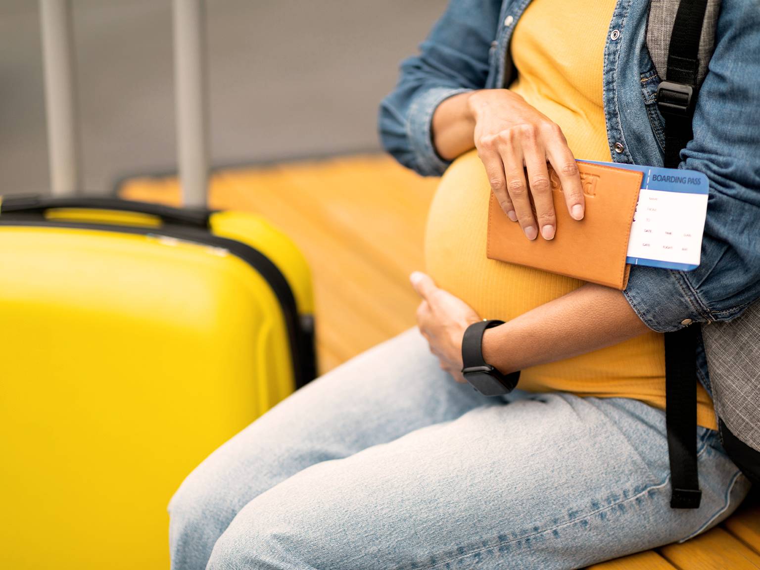 Schwangere Frau in Jeans hält einen Reisepass und eine Bordkarte und sitzt neben einem gelben Koffer.