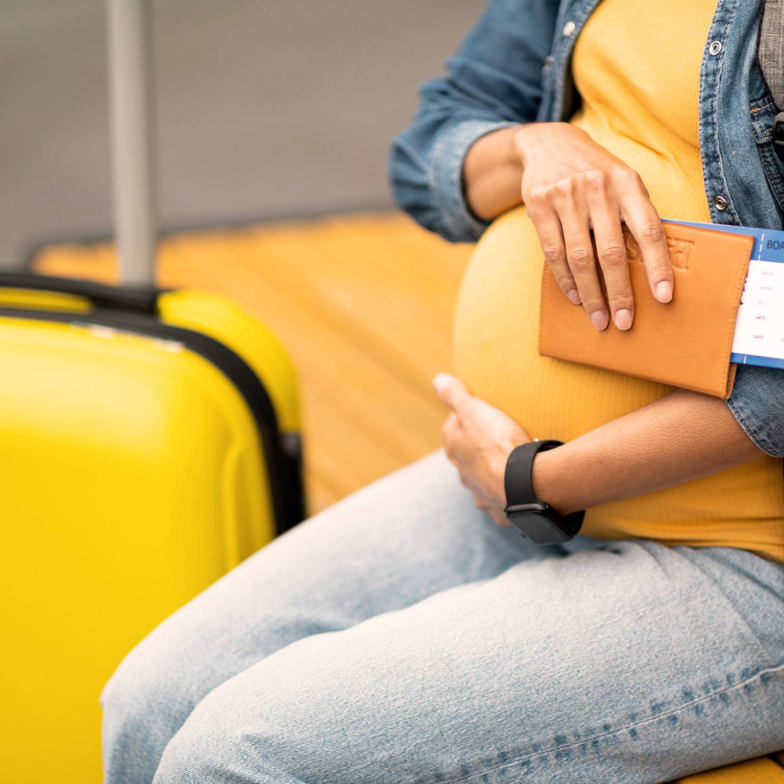 Schwangere Frau in Jeans hält einen Reisepass und eine Bordkarte und sitzt neben einem gelben Koffer.