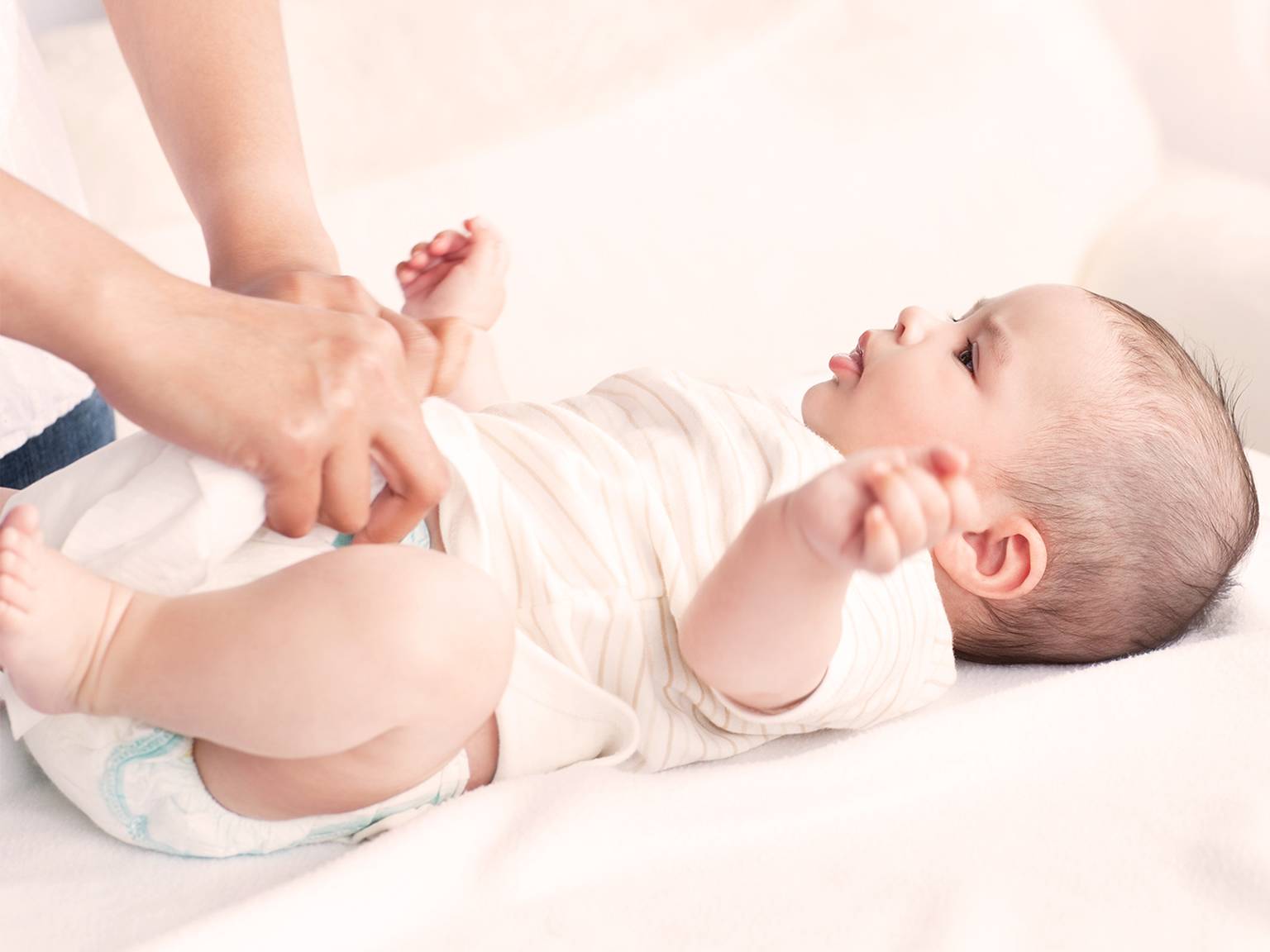 Eine Person wechselt die Windel eines Babys auf einer weichen, weißen Oberfläche. Das Baby liegt auf dem Rücken, trägt ein gestreiftes Outfit und hat die Arme leicht erhoben.