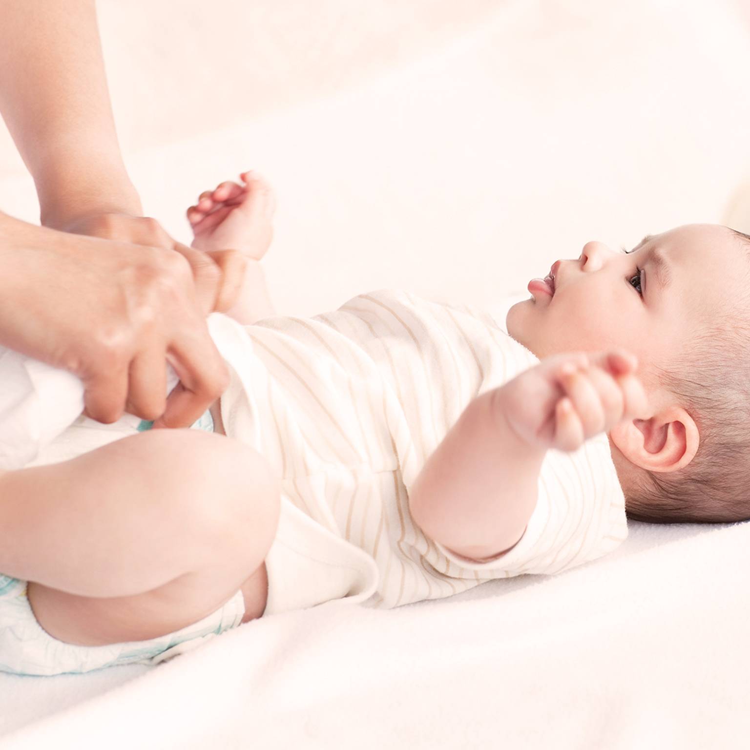 Eine Person wechselt die Windel eines Babys auf einer weichen, weißen Oberfläche. Das Baby liegt auf dem Rücken, trägt ein gestreiftes Outfit und hat die Arme leicht erhoben.