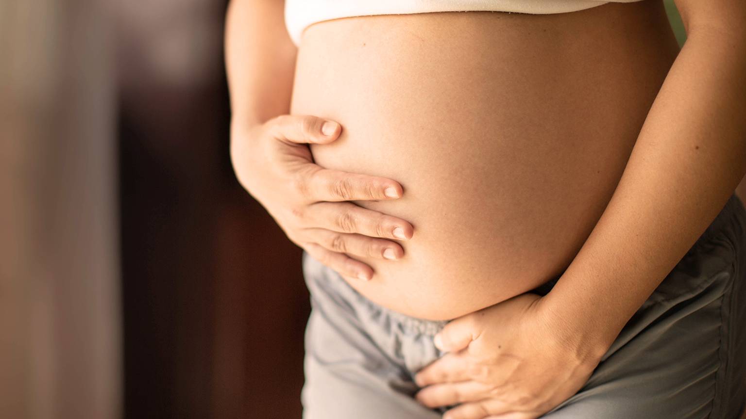 Schwangere Person in weißem Oberteil und grauen Shorts hält sanft ihren Bauch mit beiden Händen.