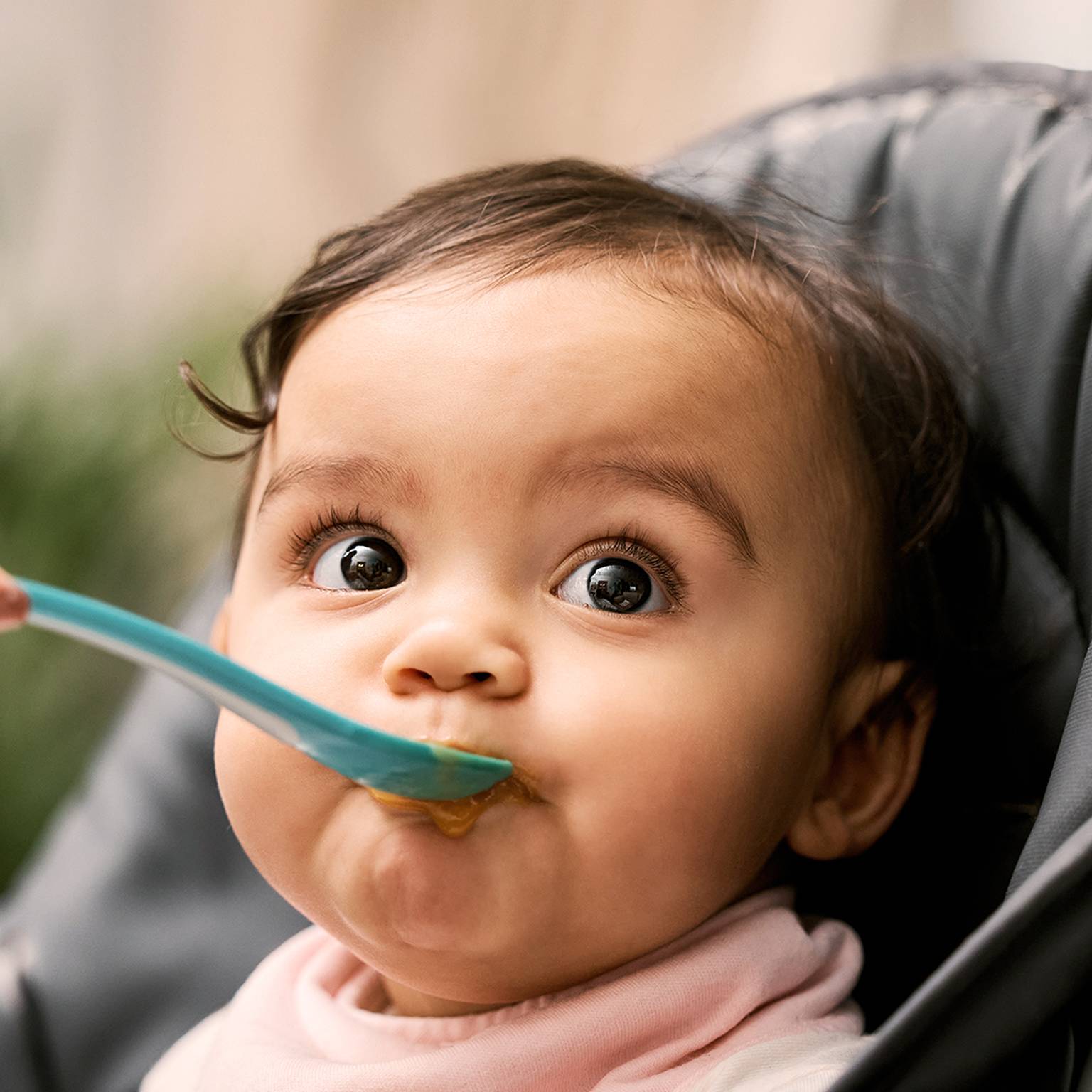 Ein Baby in einem Hochstuhl wird mit einem blauen Löffel gefüttert, hat große Augen und trägt ein rosa Lätzchen.