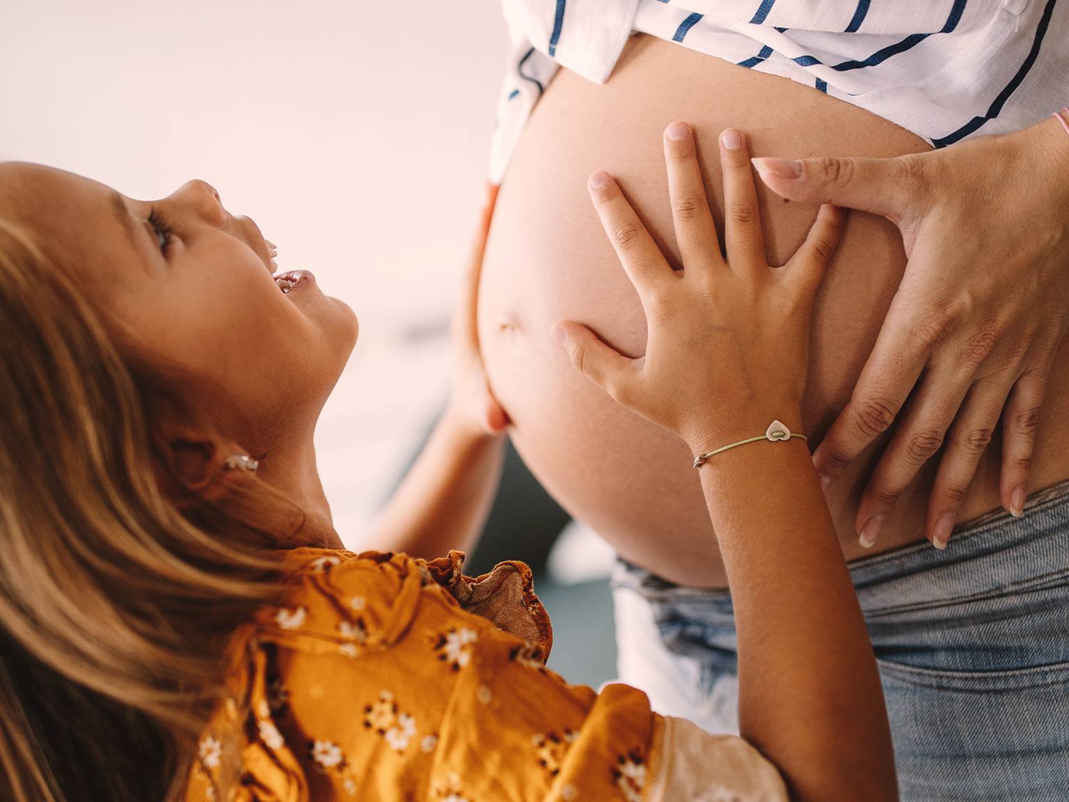 Ein lächelndes Mädchen berührt den Bauch einer schwangeren Frau und drückt damit Freude und Vorfreude aus. Die Frau trägt ein gestreiftes Hemd, das Mädchen ein geblümtes Kleid.