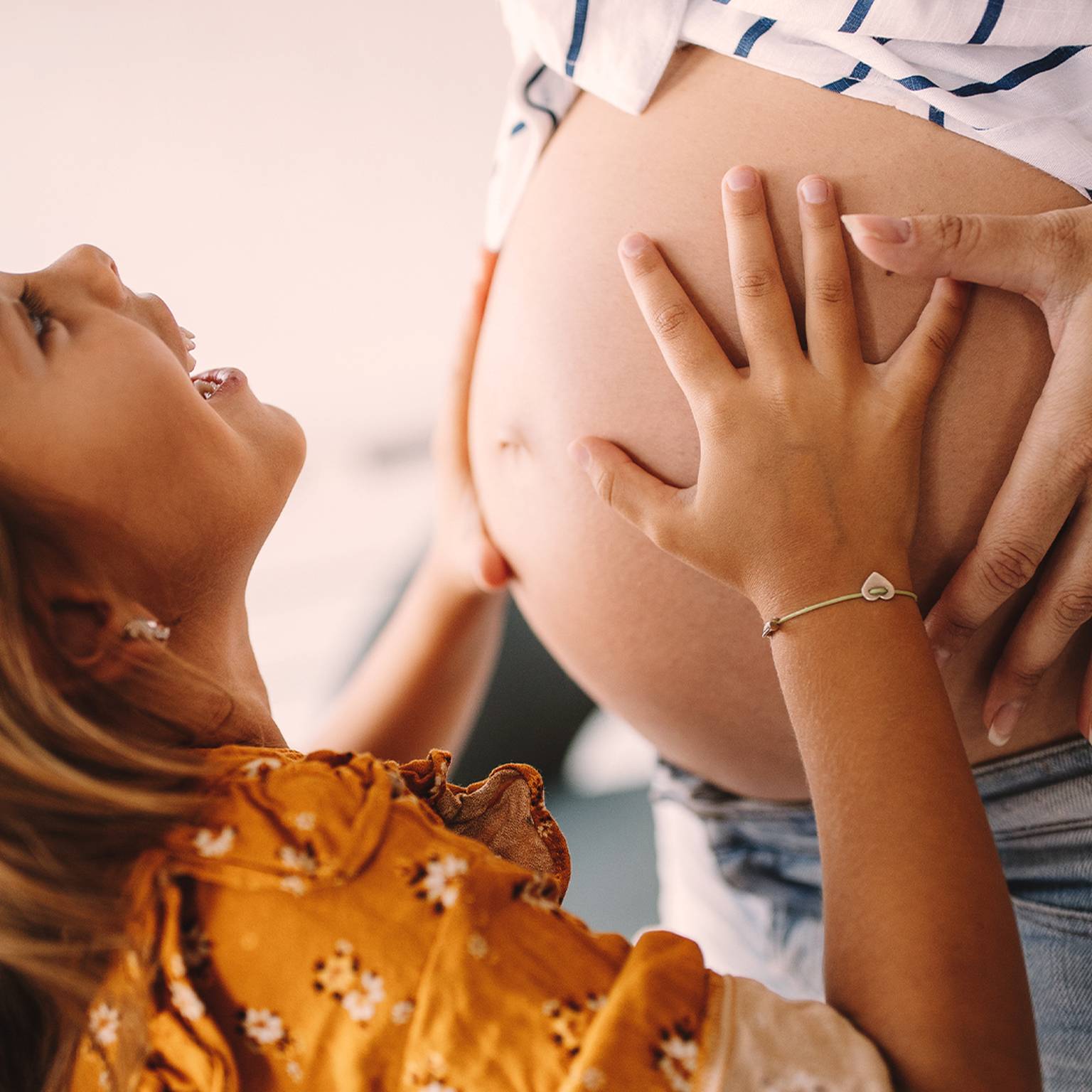 Ein lächelndes Mädchen berührt den Bauch einer schwangeren Frau und drückt damit Freude und Vorfreude aus. Die Frau trägt ein gestreiftes Hemd, das Mädchen ein geblümtes Kleid.