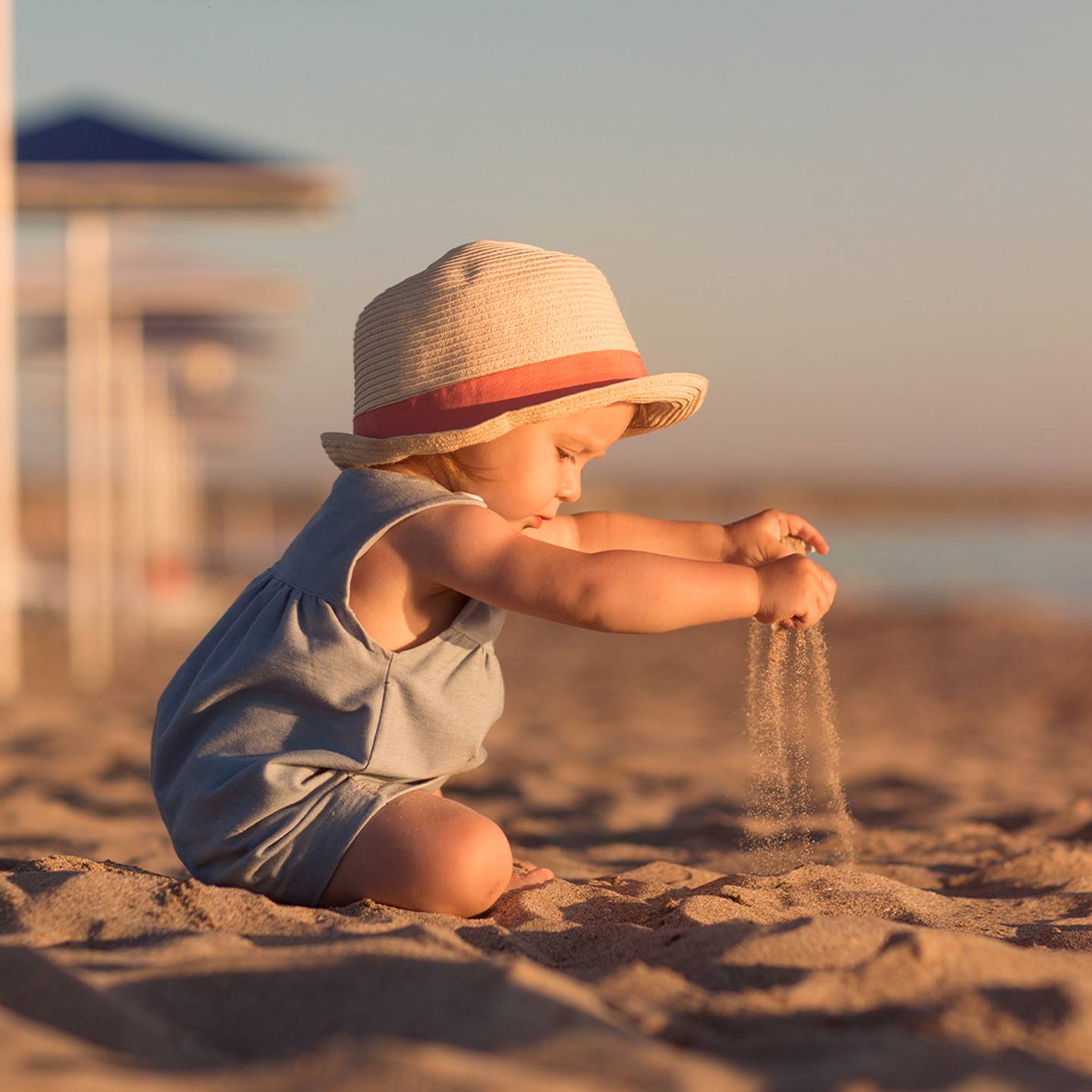 Ein Kleinkind mit Sonnenhut und blauem Outfit sitzt an einem Sandstrand und spielt unter klarem Himmel mit Sand. Im Hintergrund sind Sonnenschirme zu sehen.