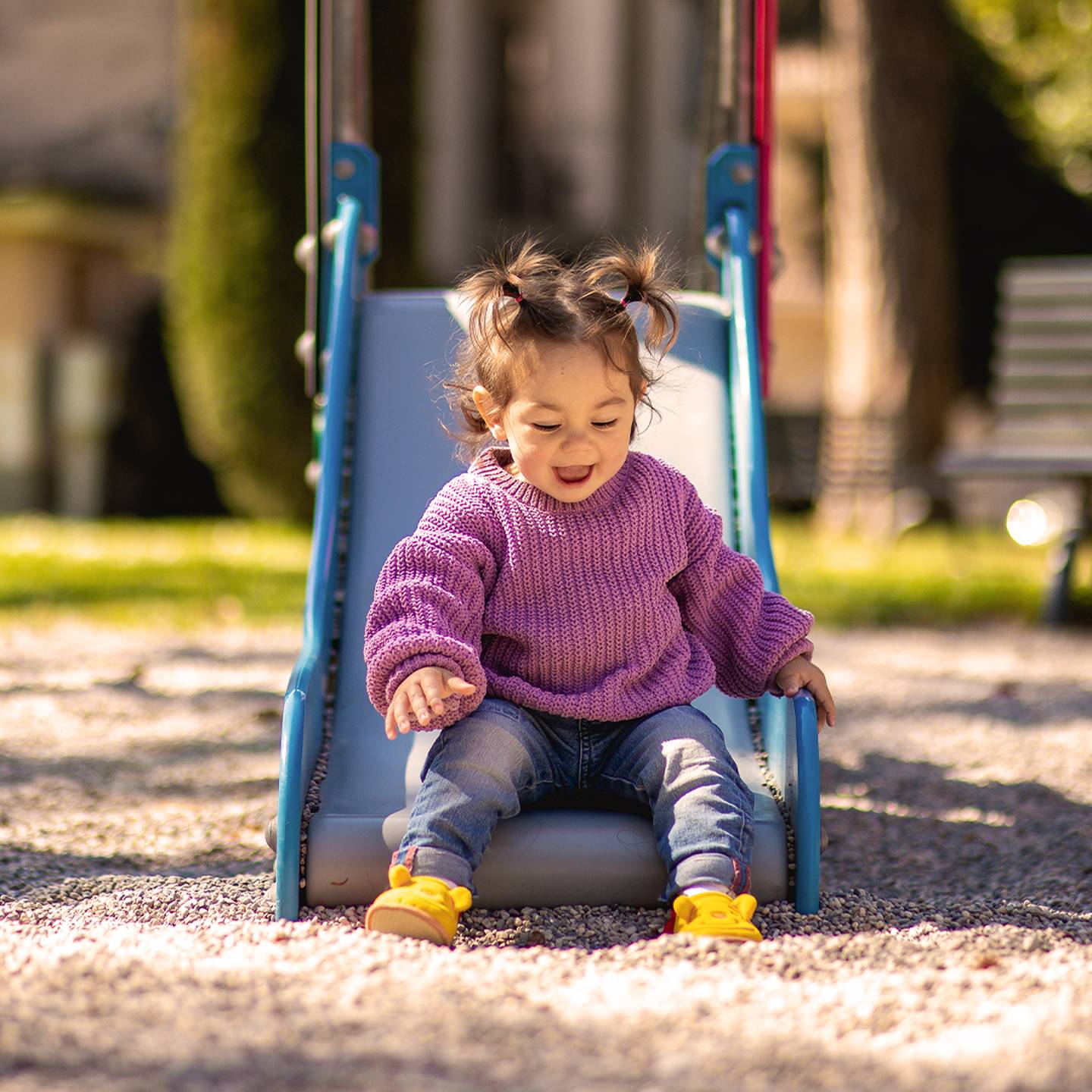 Ein Kleinkind mit Zöpfen und einem lila Pullover rutscht an einem sonnigen Tag freudig eine Rutsche auf einem Spielplatz hinunter.