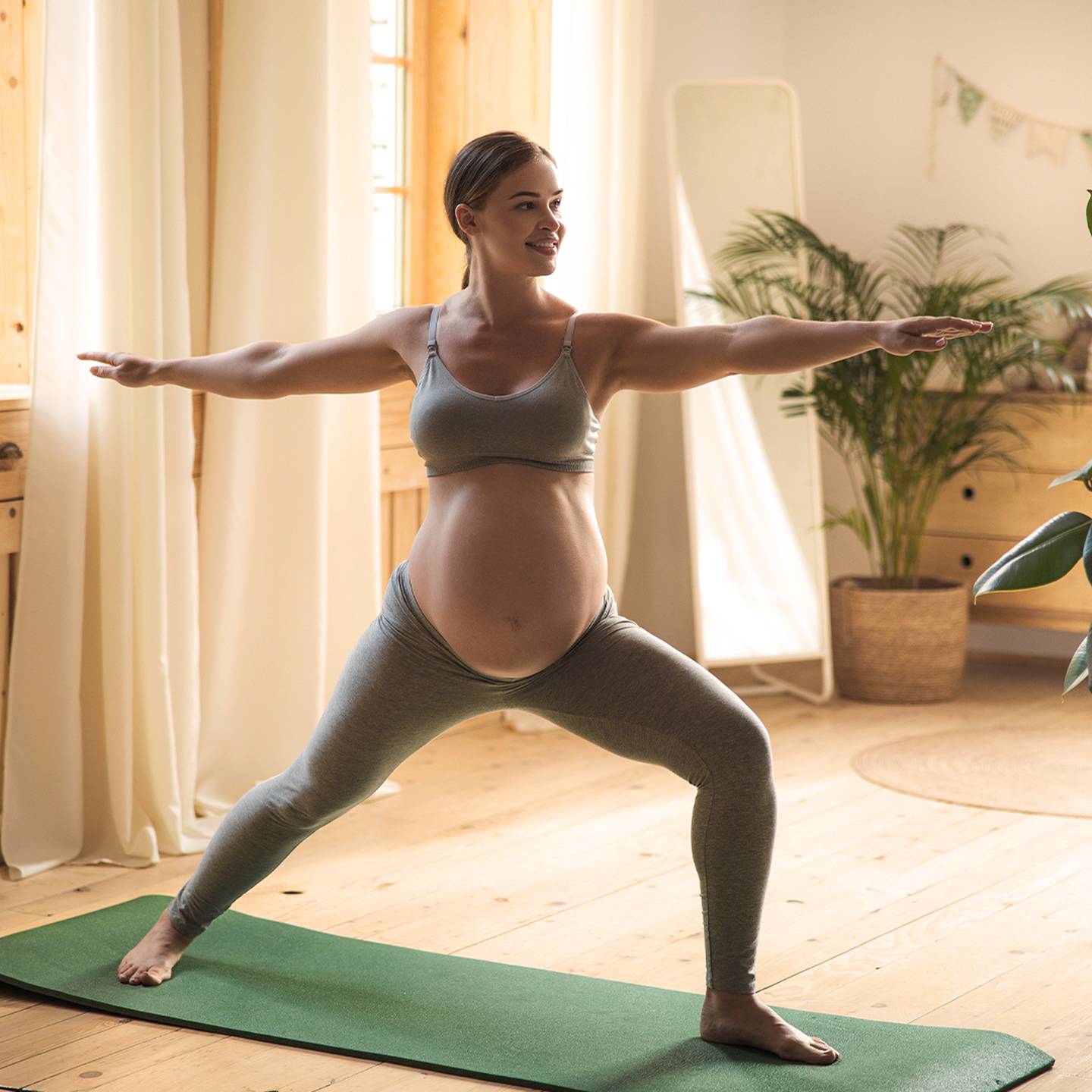 Schwangere Frau praktiziert Yoga in einem hellen Raum. Sie steht in einer Kriegerpose auf einer grünen Matte, umgeben von Pflanzen und Holzmöbeln.