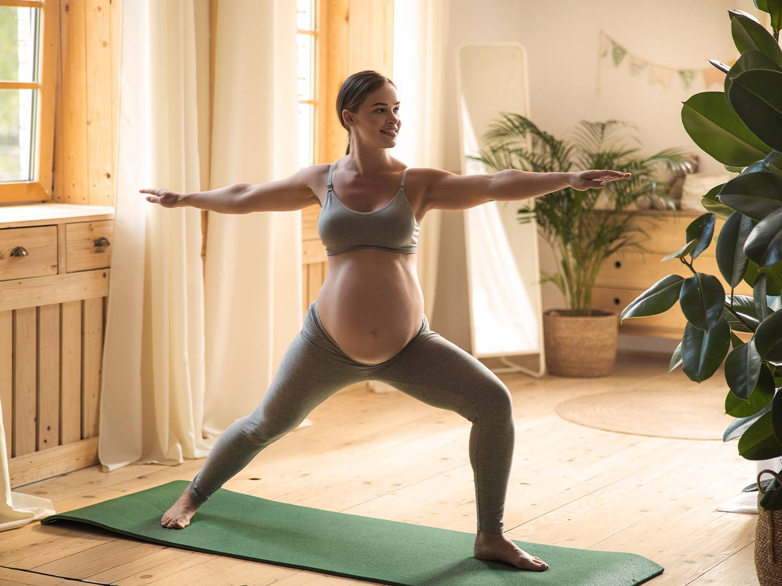 Schwangere Frau praktiziert Yoga in einem hellen Raum. Sie steht in einer Kriegerpose auf einer grünen Matte, umgeben von Pflanzen und Holzmöbeln.