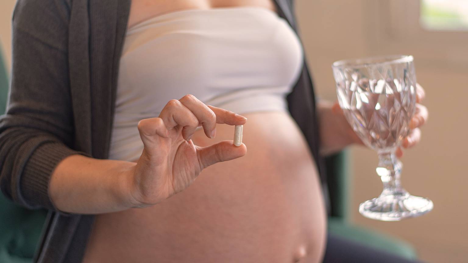 Schwangere Frau mit einer Kapsel und einem Glas Wasser in der Hand, trägt ein weißes Oberteil und eine graue Strickjacke und sitzt im Haus.