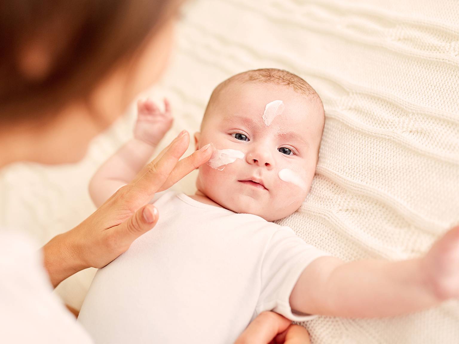 Ein Baby liegt auf einer cremefarbenen Decke, während ein Erwachsener die Wangen und die Nase des Babys eincremt.