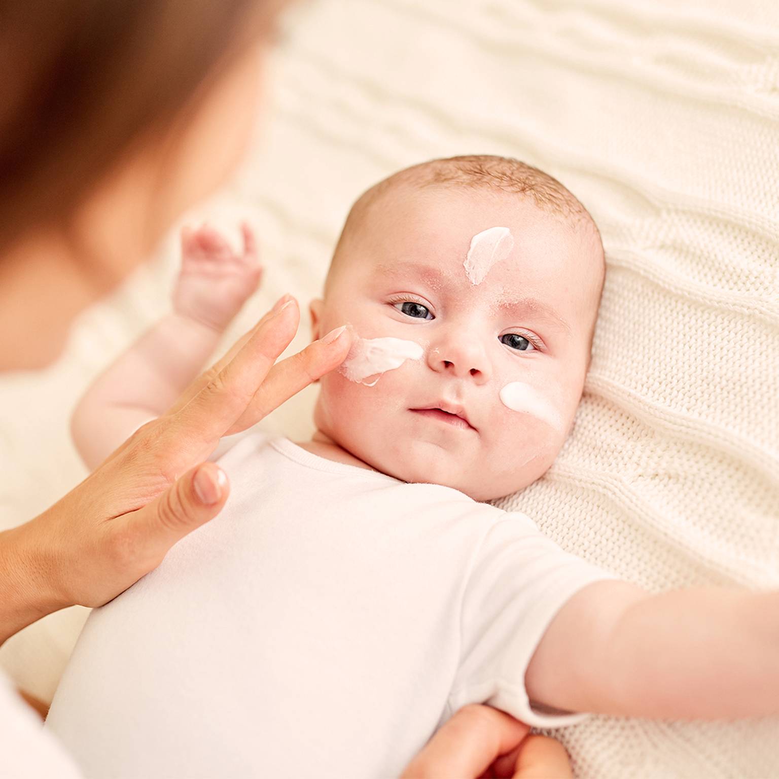 Ein Baby liegt auf einer cremefarbenen Decke, während ein Erwachsener die Wangen und die Nase des Babys eincremt.