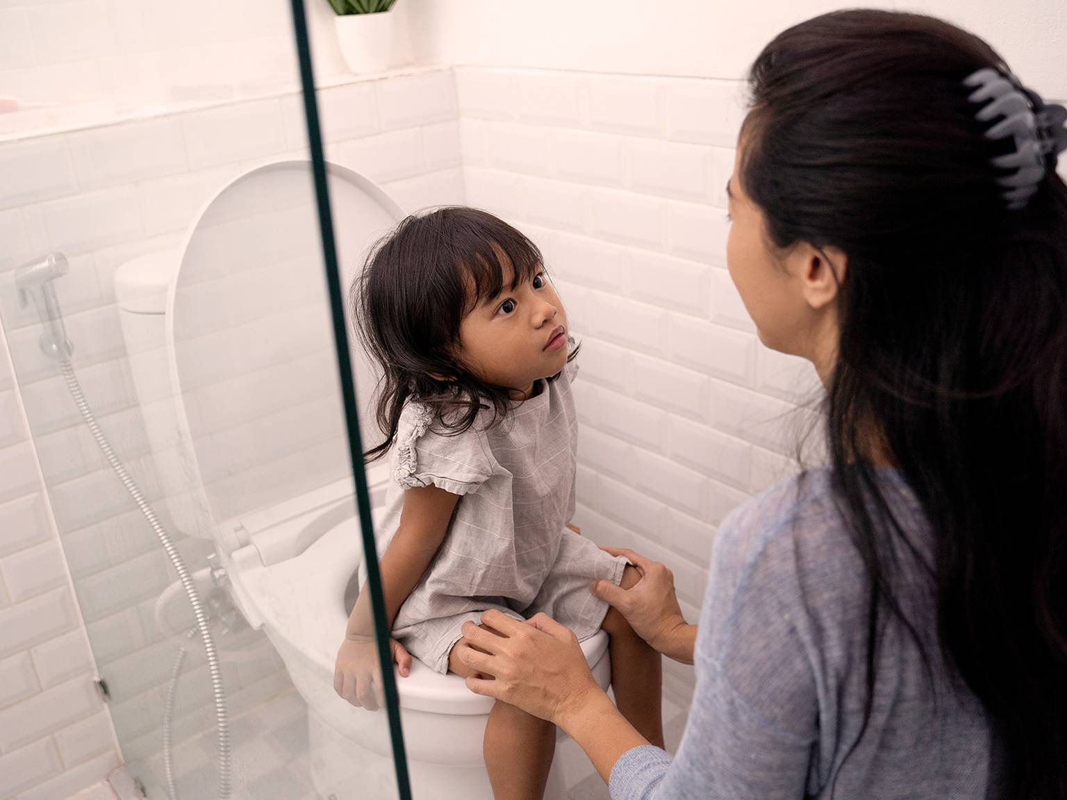 Eine Frau hilft einem kleinen Kind, das in einem Badezimmer mit weiß gefliesten Wänden auf der Toilette sitzt, und hält dabei die Hände des Kindes fest.