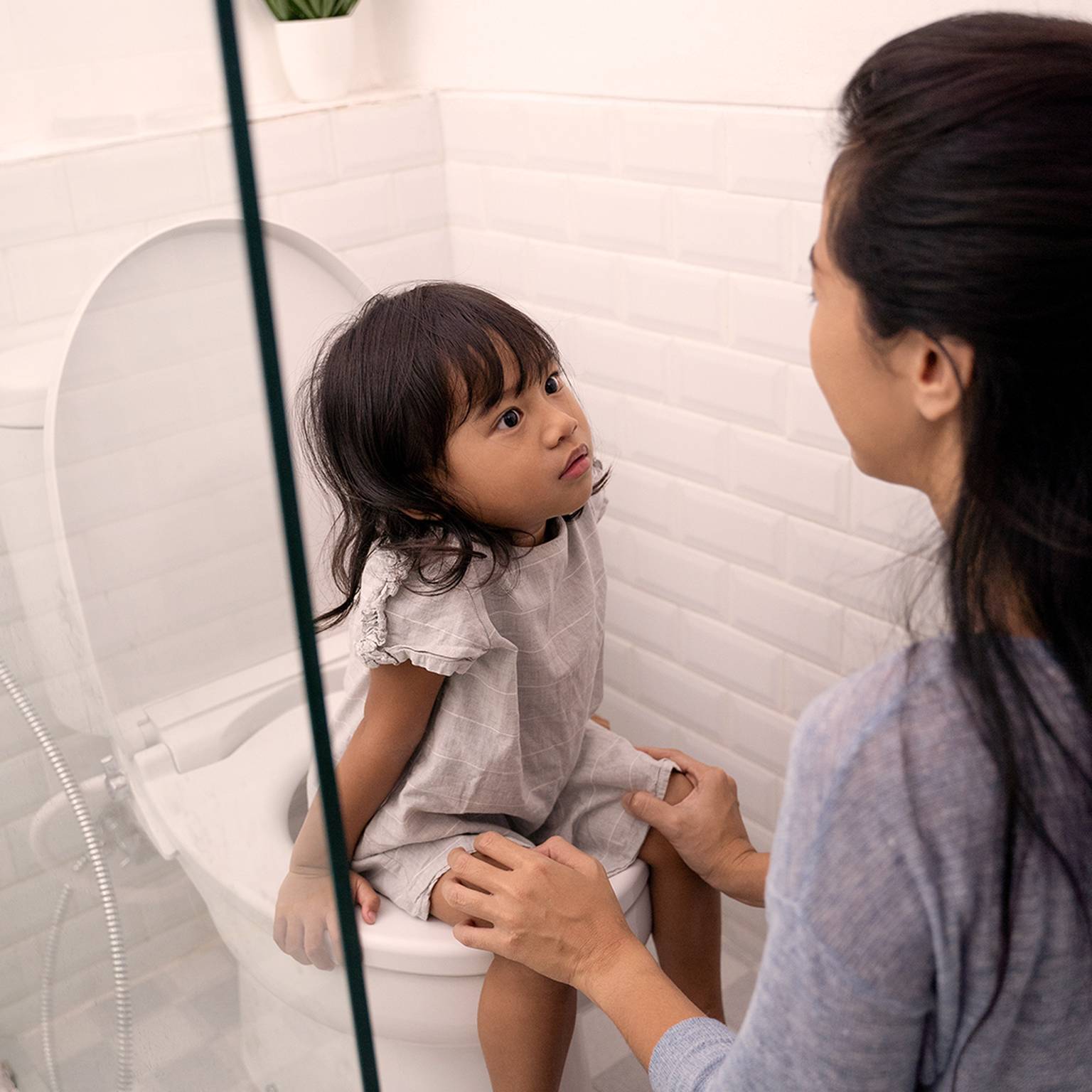 Eine Frau hilft einem kleinen Kind, das in einem Badezimmer mit weiß gefliesten Wänden auf der Toilette sitzt, und hält dabei die Hände des Kindes fest.
