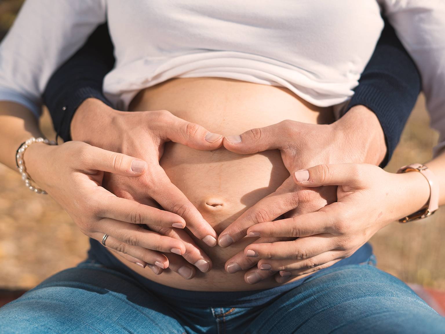 Schwangere Frau mit den Händen auf dem Bauch, die eine Herzform bilden, trägt ein weißes Oberteil und blaue Jeans, im Freien.