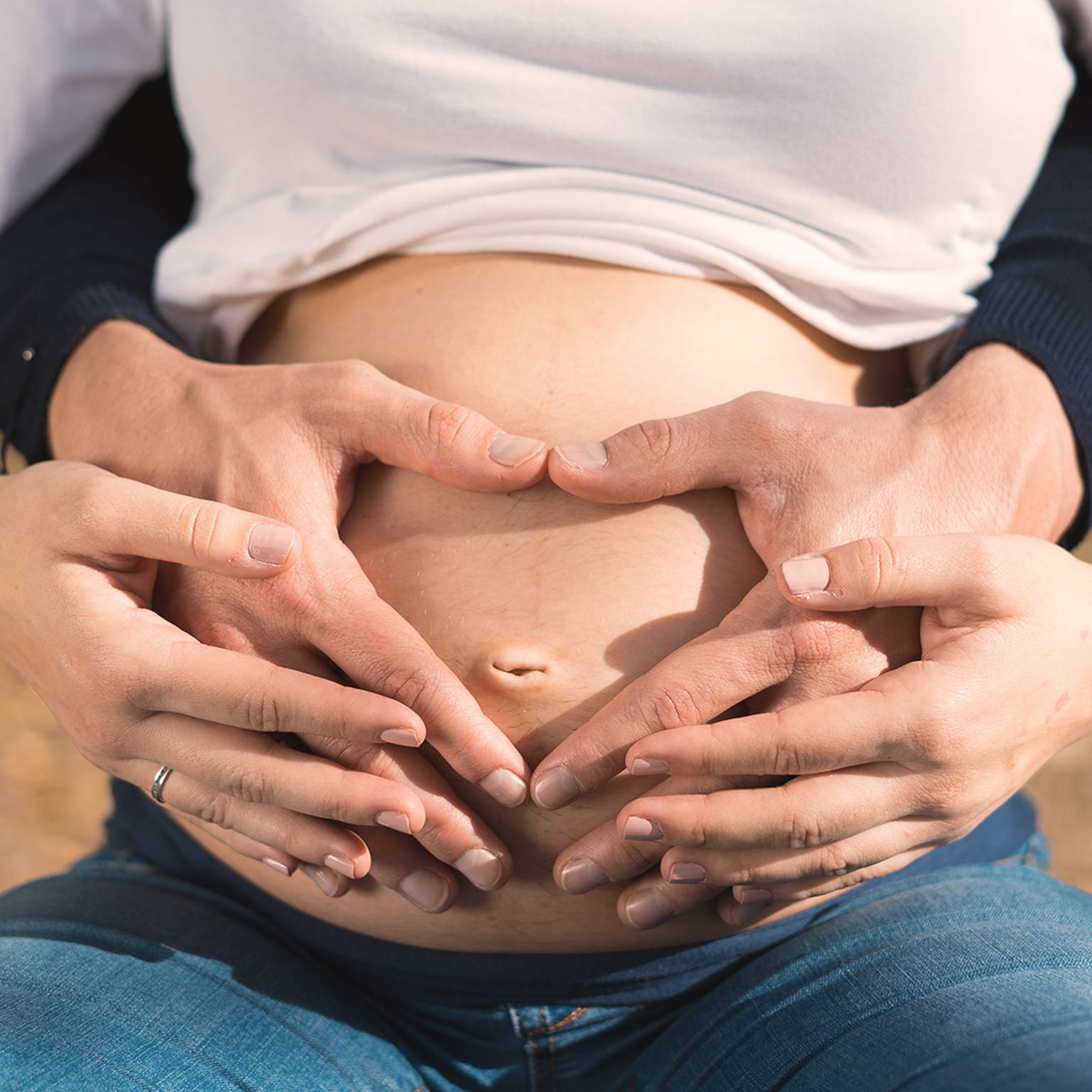 Schwangere Frau mit den Händen auf dem Bauch, die eine Herzform bilden, trägt ein weißes Oberteil und blaue Jeans, im Freien.
