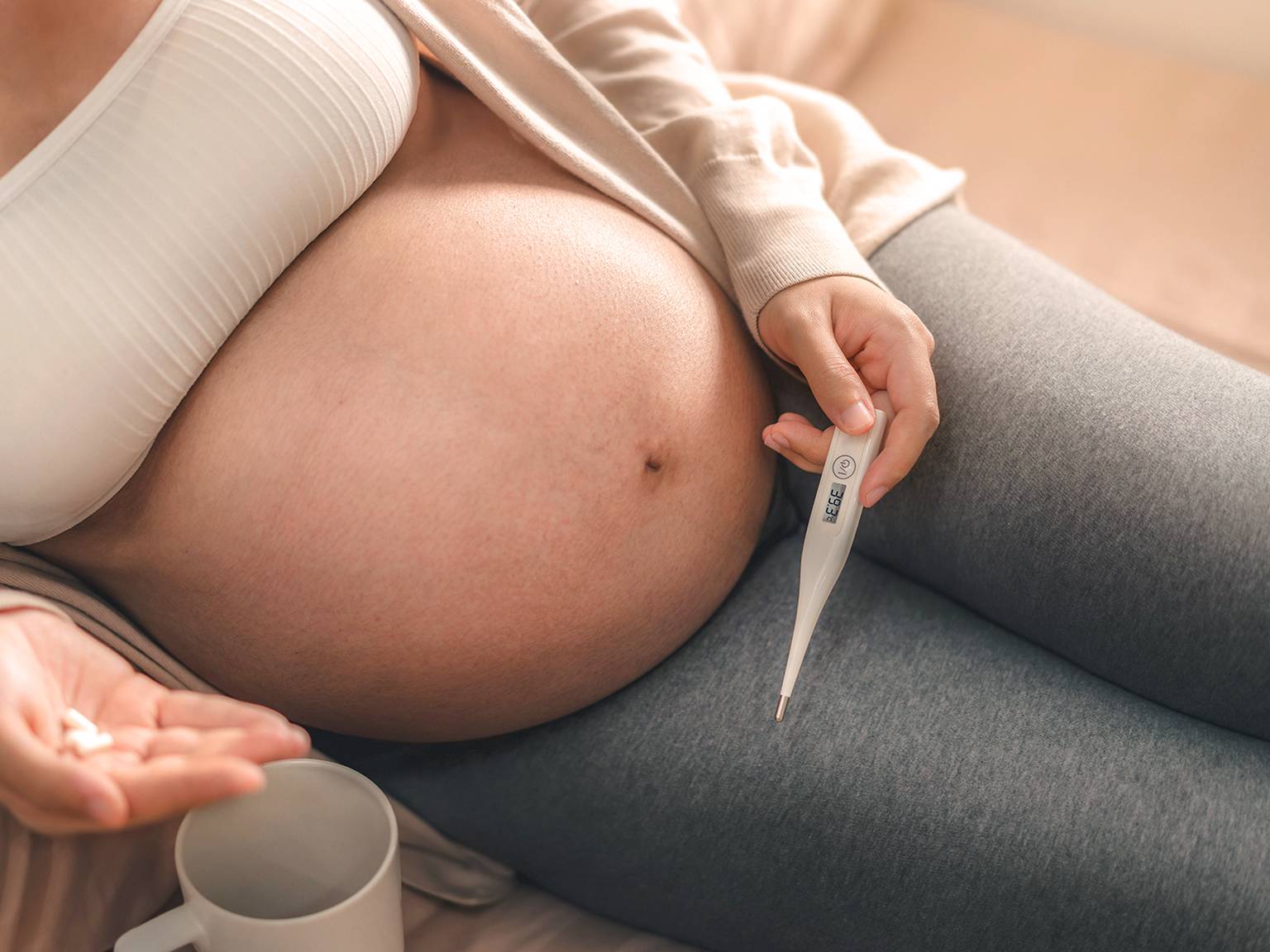 Schwangere Frau in weißem Oberteil und grauen Leggings, hält ein Thermometer in der Hand und sitzt auf einem Bett mit einer Tasse daneben.
