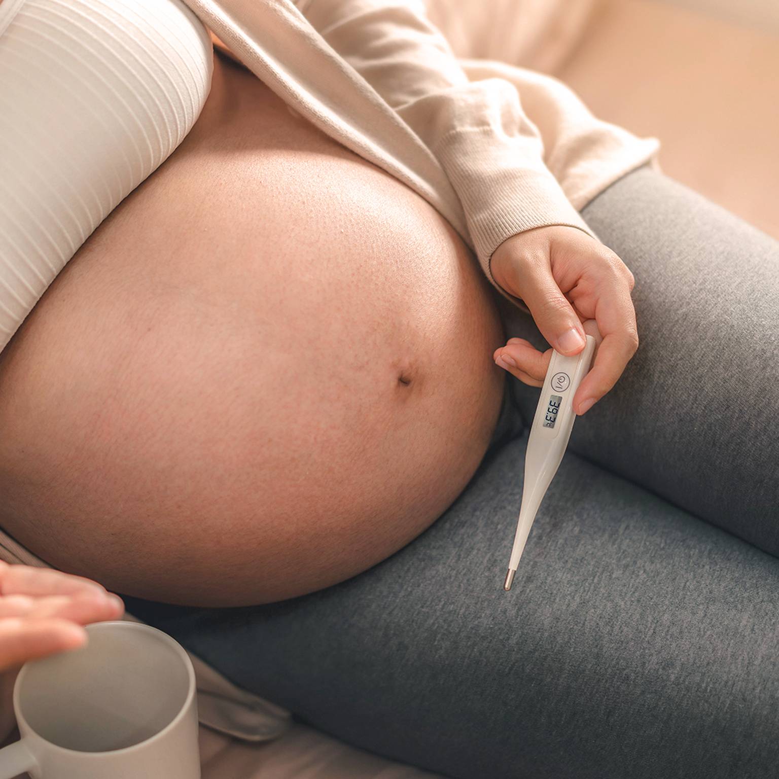 Schwangere Frau in weißem Oberteil und grauen Leggings, hält ein Thermometer in der Hand und sitzt auf einem Bett mit einer Tasse daneben.