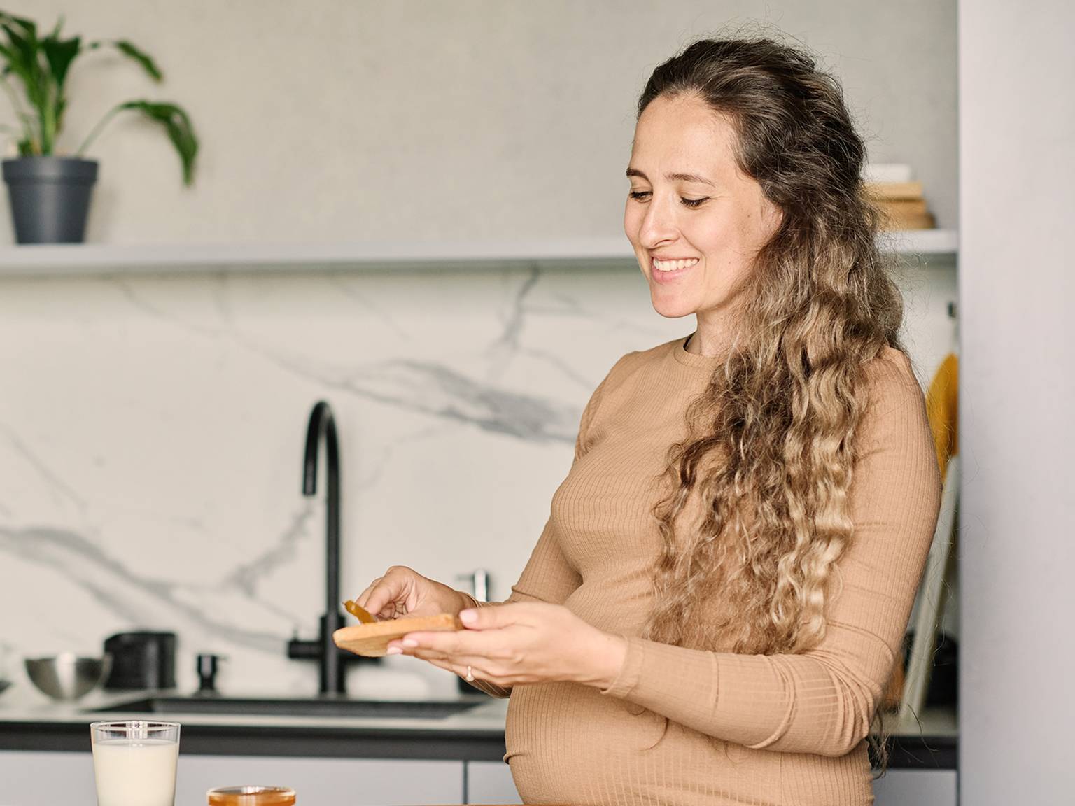 Lächelnde Frau mit langem lockigem Haar in einem hellbraunen Pullover hält ein Stück Brot in einer modernen Küche mit Marmorrückwand und schwarzem Wasserhahn.