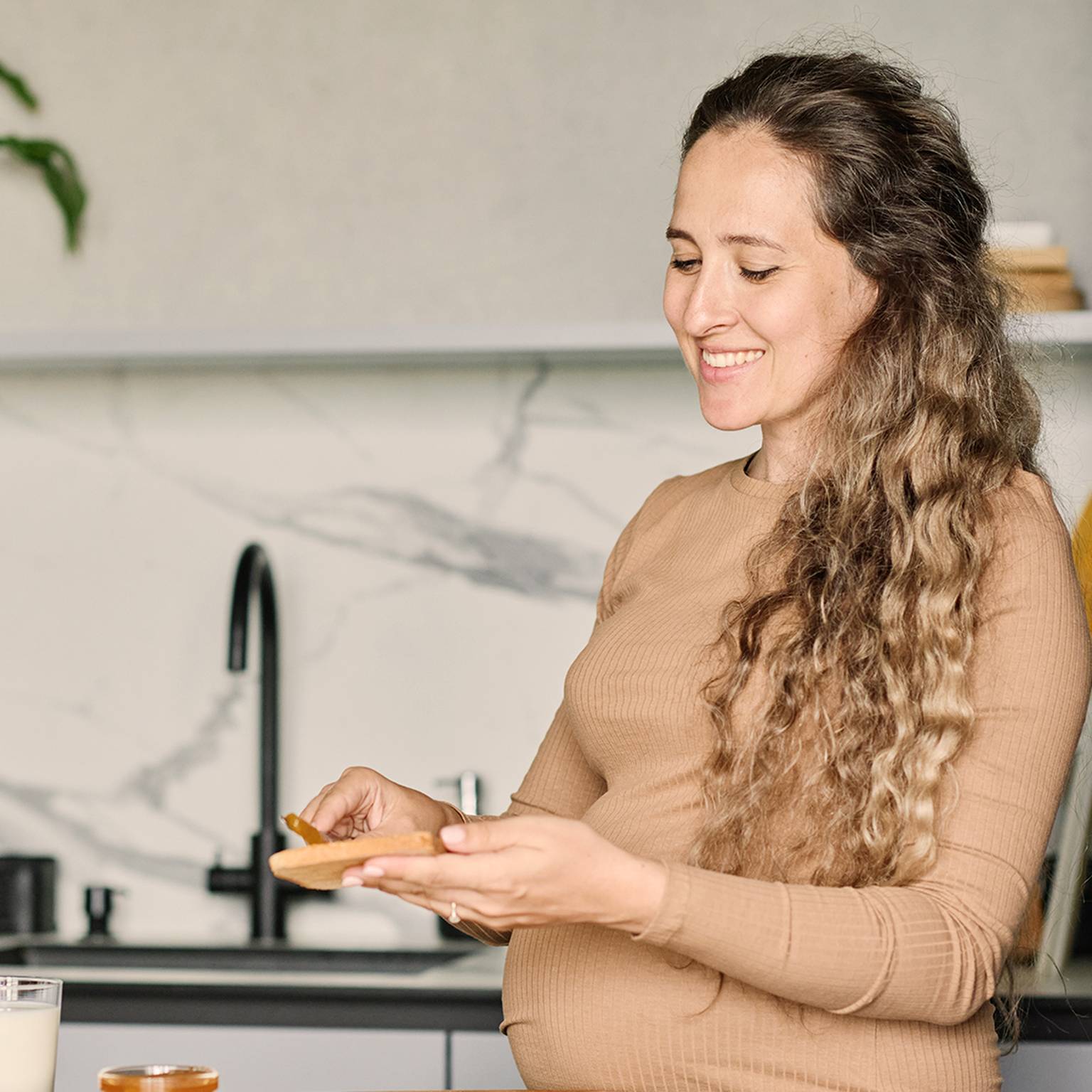 Lächelnde Frau mit langem lockigem Haar in einem hellbraunen Pullover hält ein Stück Brot in einer modernen Küche mit Marmorrückwand und schwarzem Wasserhahn.