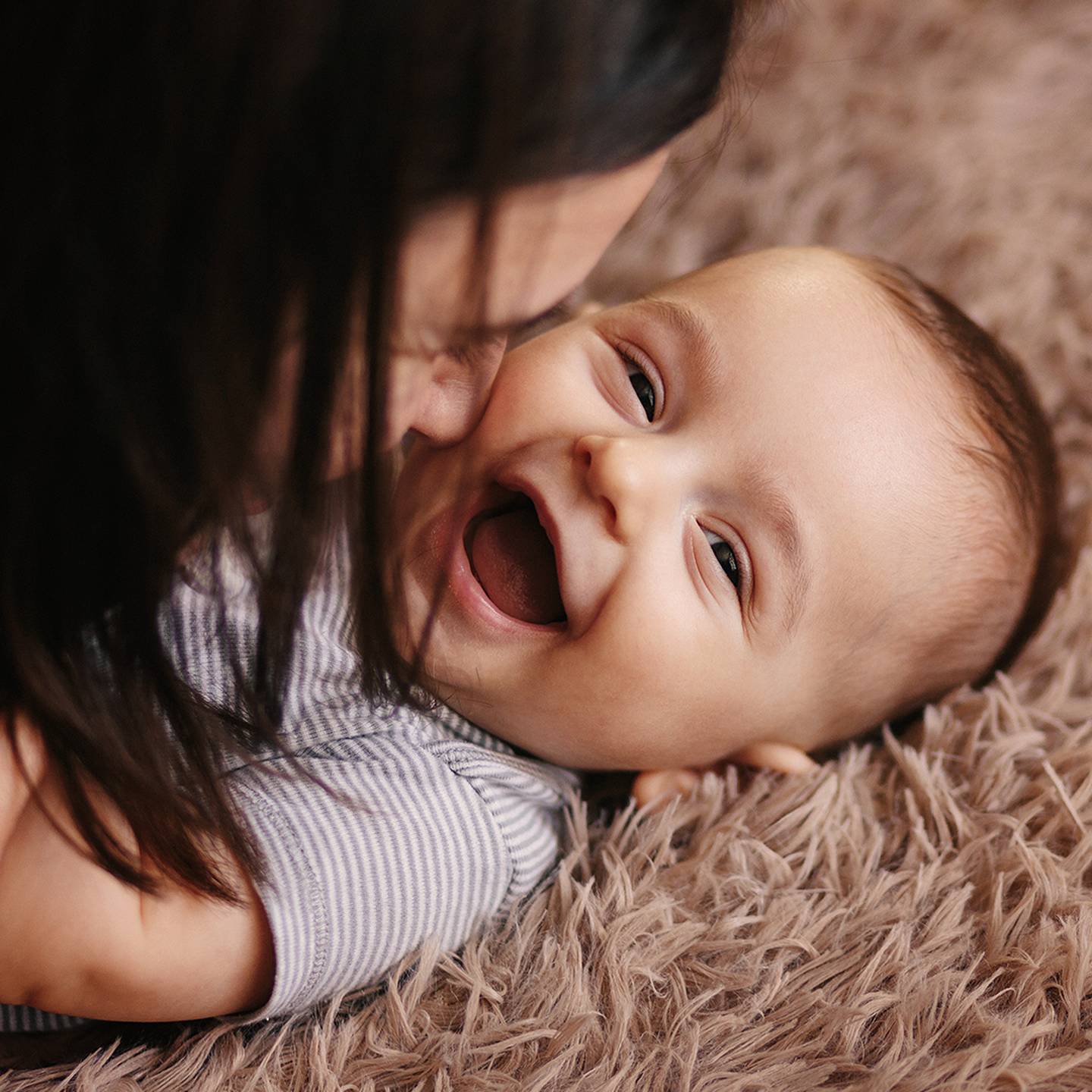 Ein lächelndes Baby liegt auf einer weichen, pelzigen Oberfläche und interagiert spielerisch mit einer Person, deren Gesicht dem des Babys nahe ist.