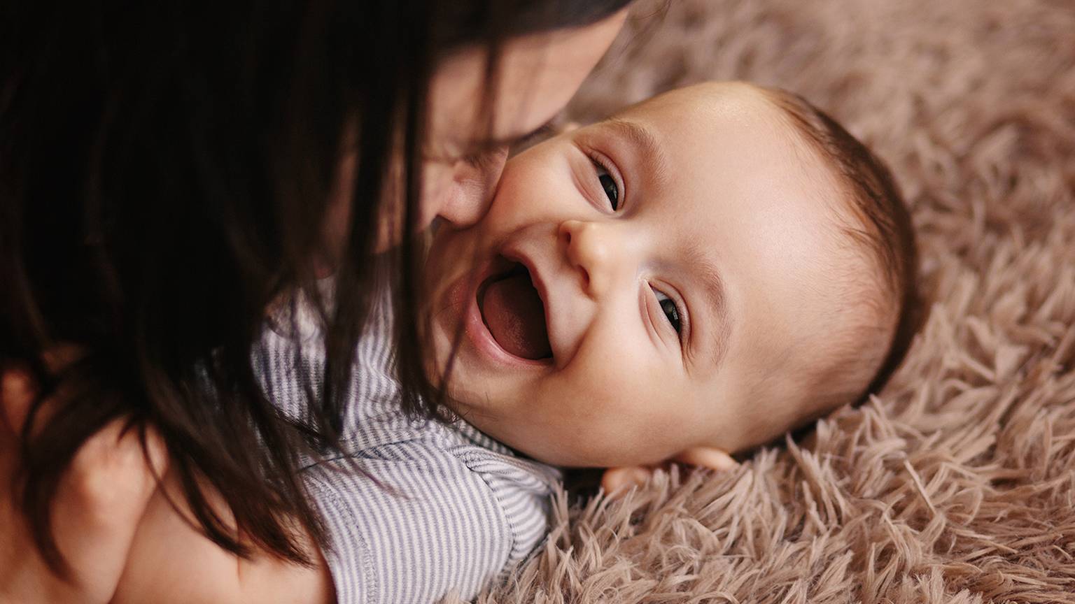 Ein lächelndes Baby liegt auf einer weichen, pelzigen Oberfläche und interagiert spielerisch mit einer Person, deren Gesicht dem des Babys nahe ist.