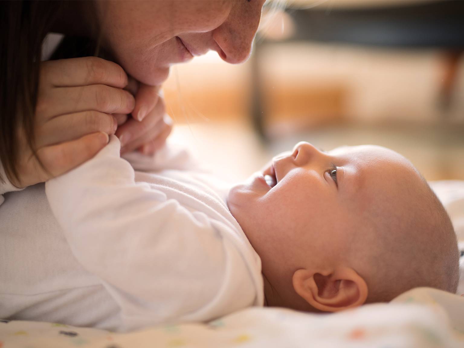 Eine Frau hält sanft die Hände eines lächelnden Babys, das auf einer weichen Decke liegt und einen zärtlichen Moment teilt.