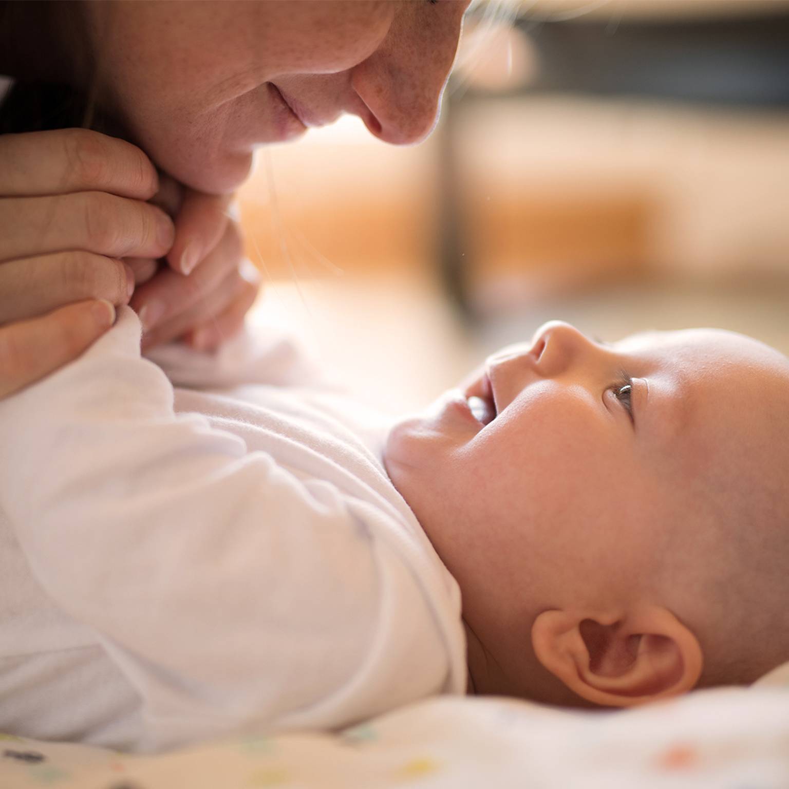 Eine Frau hält sanft die Hände eines lächelnden Babys, das auf einer weichen Decke liegt und einen zärtlichen Moment teilt.