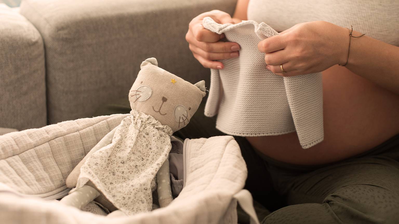 Eine schwangere Frau faltet Babykleidung neben einem Korb mit einem Kuscheltier und sitzt auf einem Sofa.