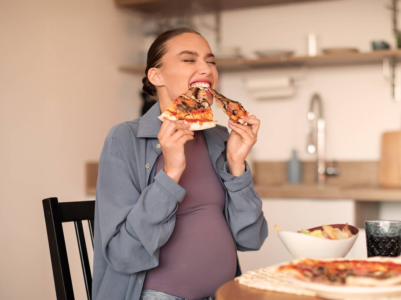Schwangere Frau isst freudig zwei Stücke Pizza an einem Küchentisch, daneben steht eine Salatschüssel.