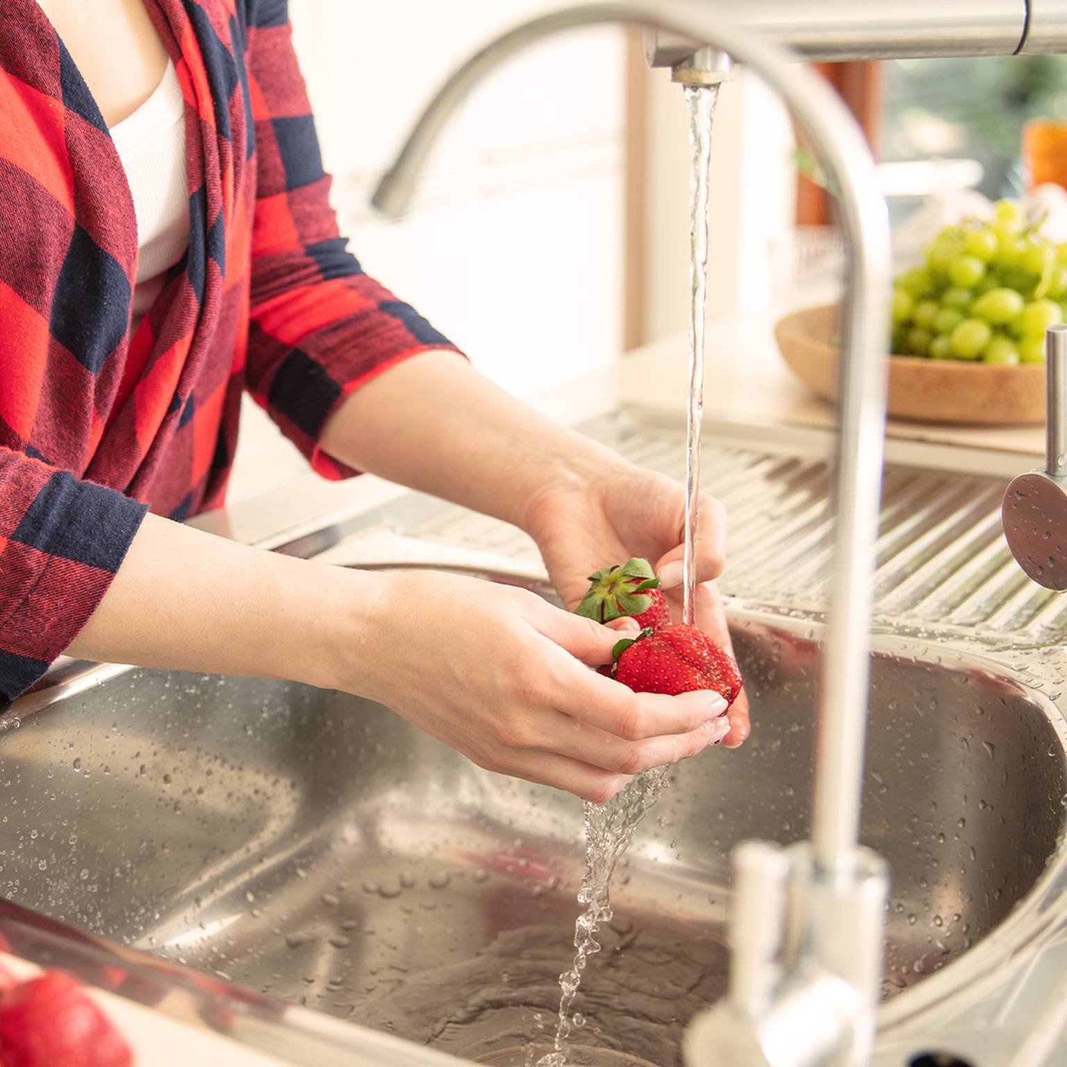 Person im karierten Hemd wäscht Erdbeeren unter einem Küchenwasserhahn, auf der Arbeitsplatte liegt noch mehr Obst.