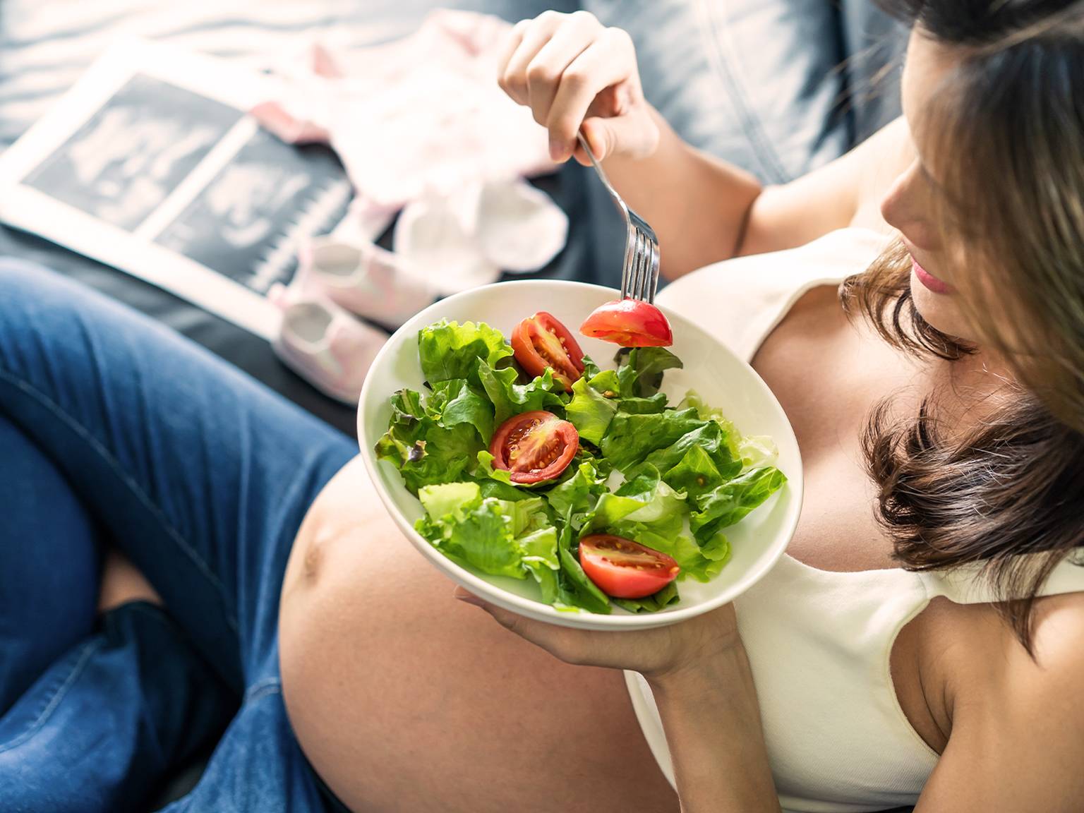 Schwangere Frau sitzt auf einer Couch und isst einen Salat mit Tomaten und Gemüse. Im Hintergrund sind Babykleidung und Schuhe zu sehen.