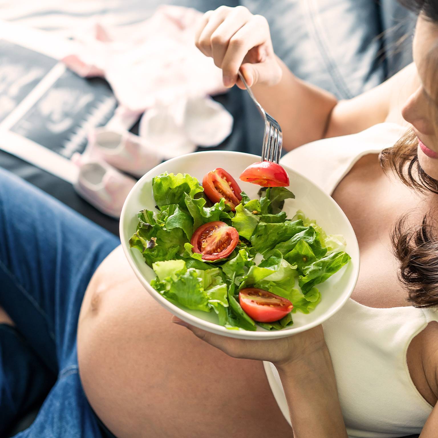 Schwangere Frau sitzt auf einer Couch und isst einen Salat mit Tomaten und Gemüse. Im Hintergrund sind Babykleidung und Schuhe zu sehen.