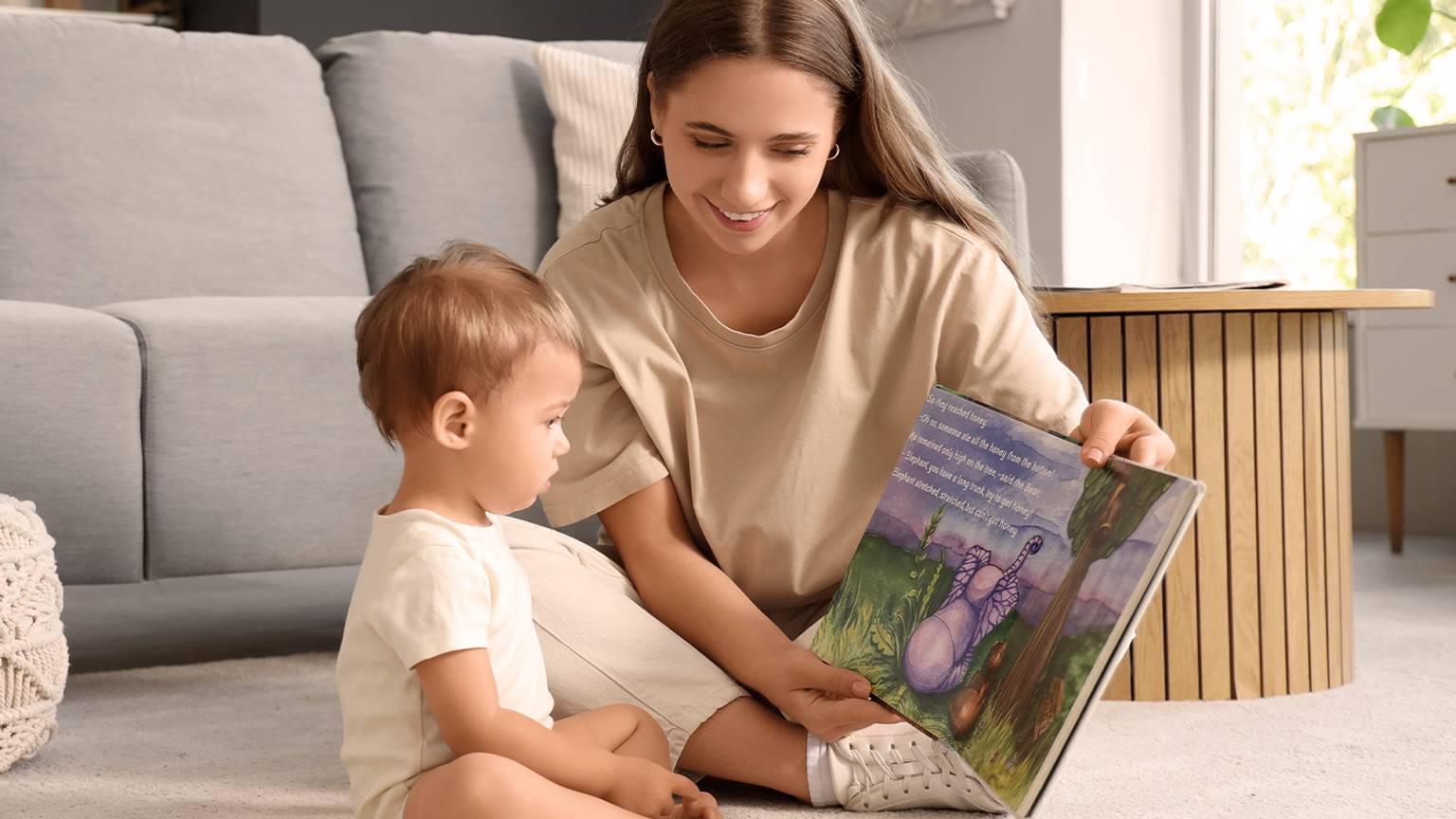 In einem gemütlichen Wohnzimmer mit einem grauen Sofa und einem runden Holztisch liest eine Frau einem Kleinkind auf dem Boden ein Kinderbuch vor.