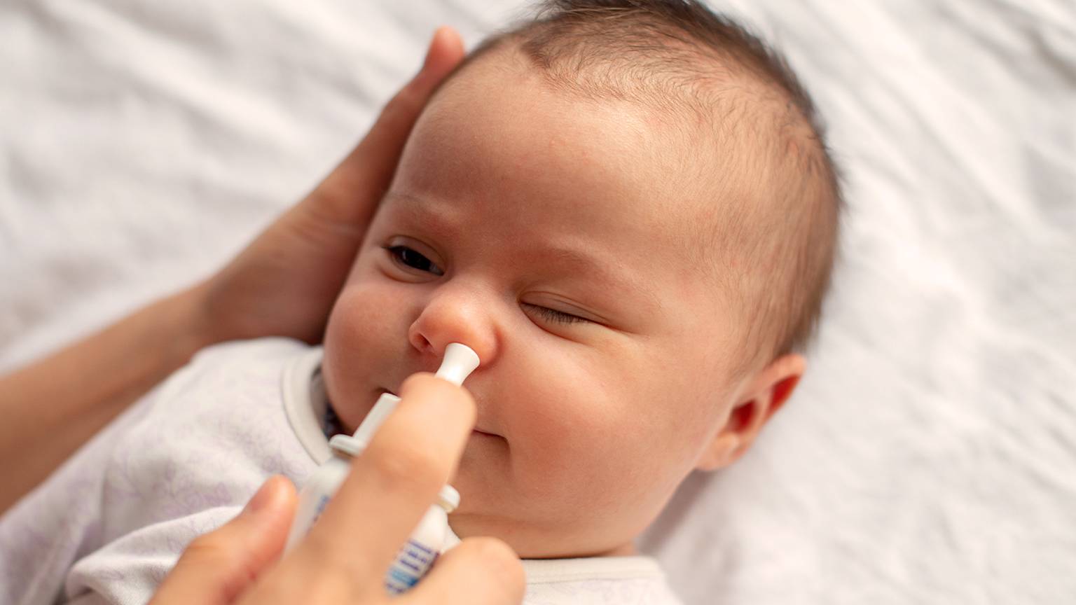 Ein Baby liegt auf einer weißen Decke und bekommt von der Hand einer Pflegekraft ein Nasenspray eingesprüht. Ein Auge ist sanft geschlossen.