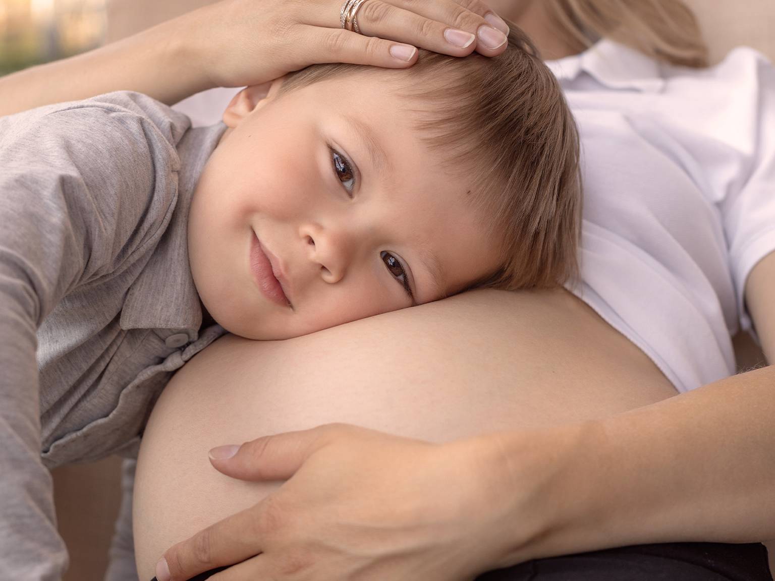 Kleiner Junge liegt fröhlich mit dem Kopf auf dem schwangeren Bauch seiner Mutter