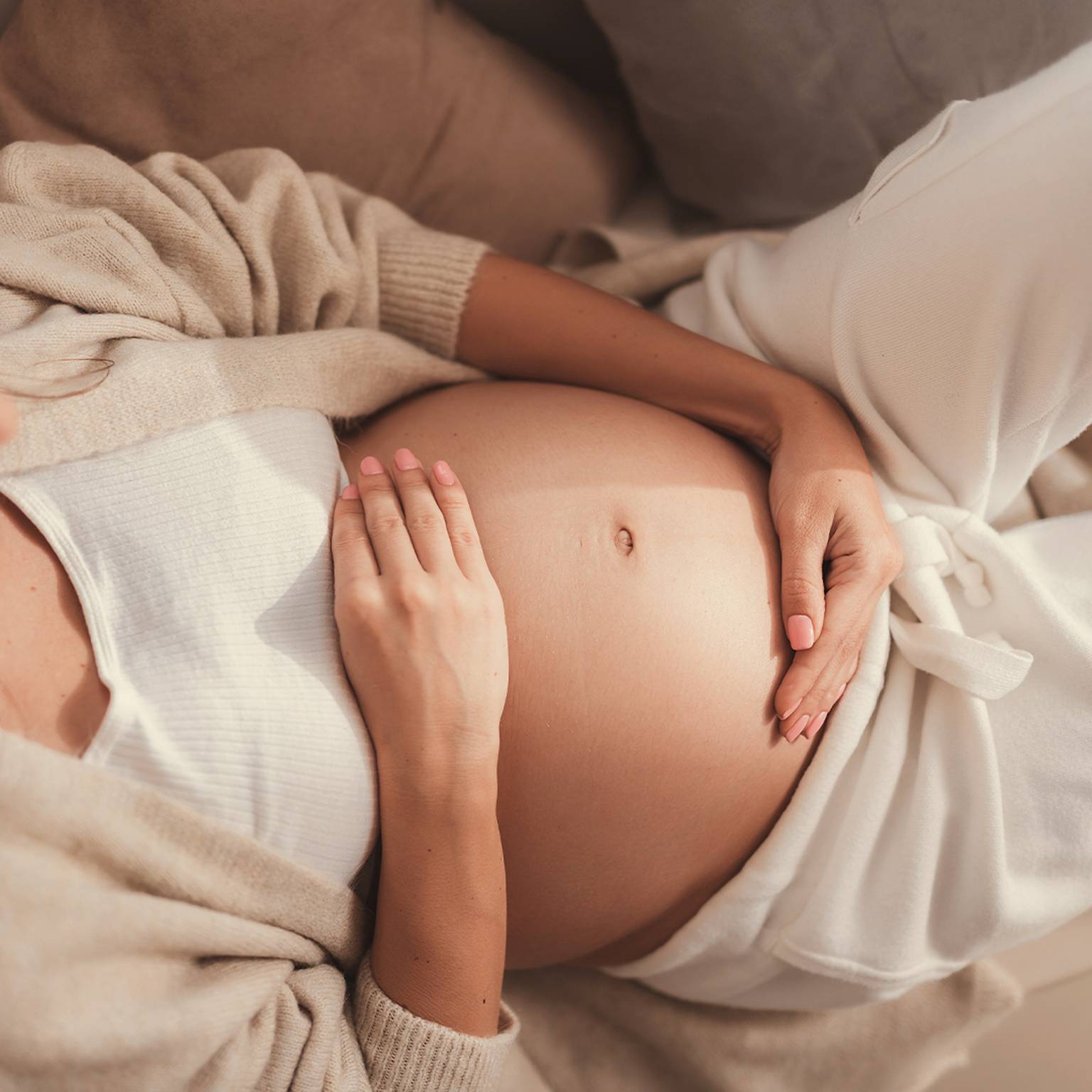 Schwangere Frau in gemütlicher Kleidung berührt sanft ihren Bauch, während sie auf einem Sofa liegt, und vermittelt so Wärme und Geborgenheit.
