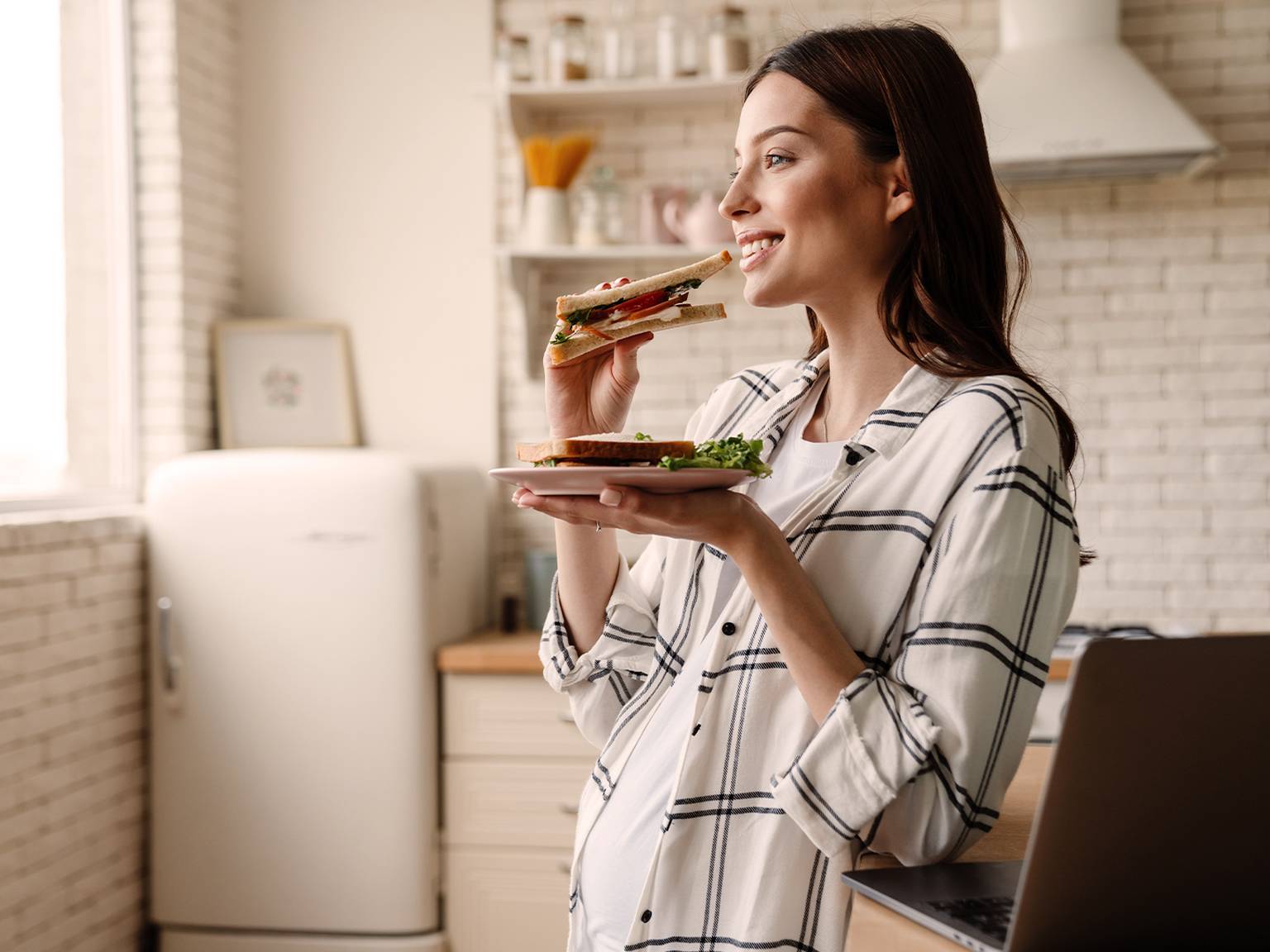 Eine Frau in einem karierten Hemd lächelt, während sie in einer hellen Küche ein Sandwich isst.