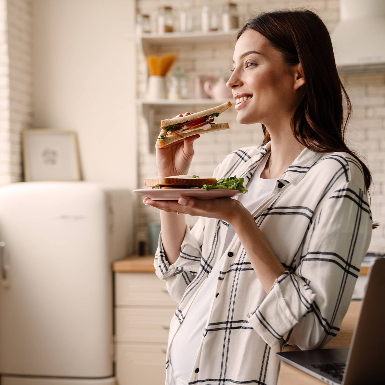 Eine Frau in einem karierten Hemd lächelt, während sie in einer hellen Küche ein Sandwich isst.