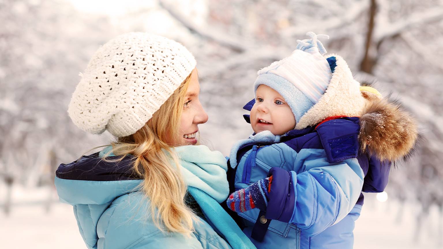 Eine Frau mit weißer Strickmütze hält ein lächelndes Baby in einem blauen Schneeanzug, umgeben von einer verschneiten Winterlandschaft.