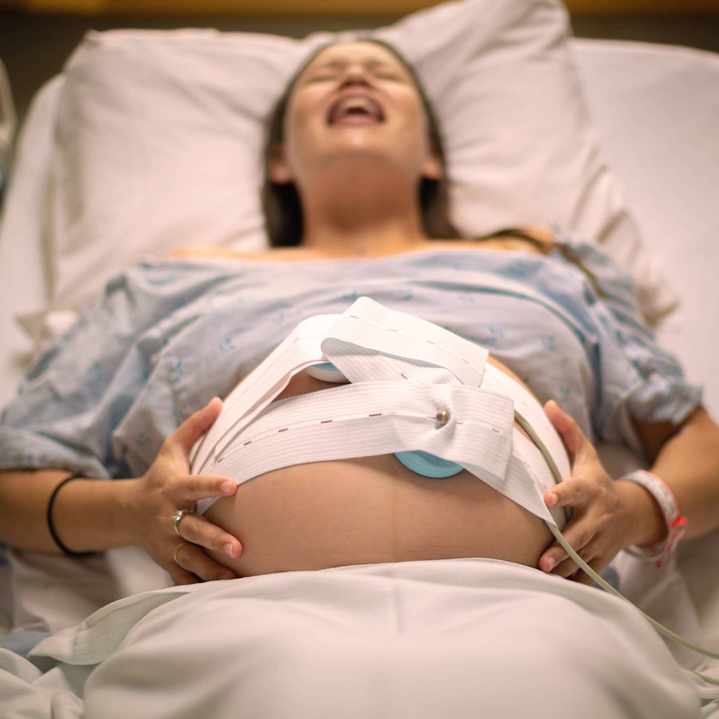 Schwangere Frau im Krankenhausbett, trägt ein Krankenhaushemd, hat Wehen und hat ein Gerät zur fetalen Überwachung auf ihrem Bauch.