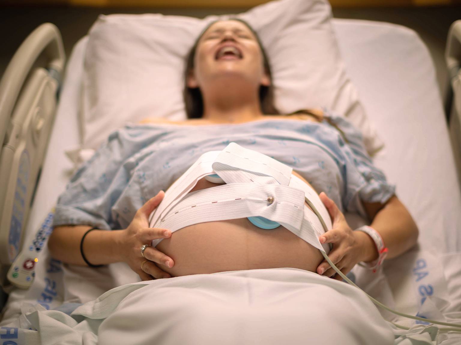 Schwangere Frau im Krankenhausbett, trägt ein Krankenhaushemd, hat Wehen und hat ein Gerät zur fetalen Überwachung auf ihrem Bauch.