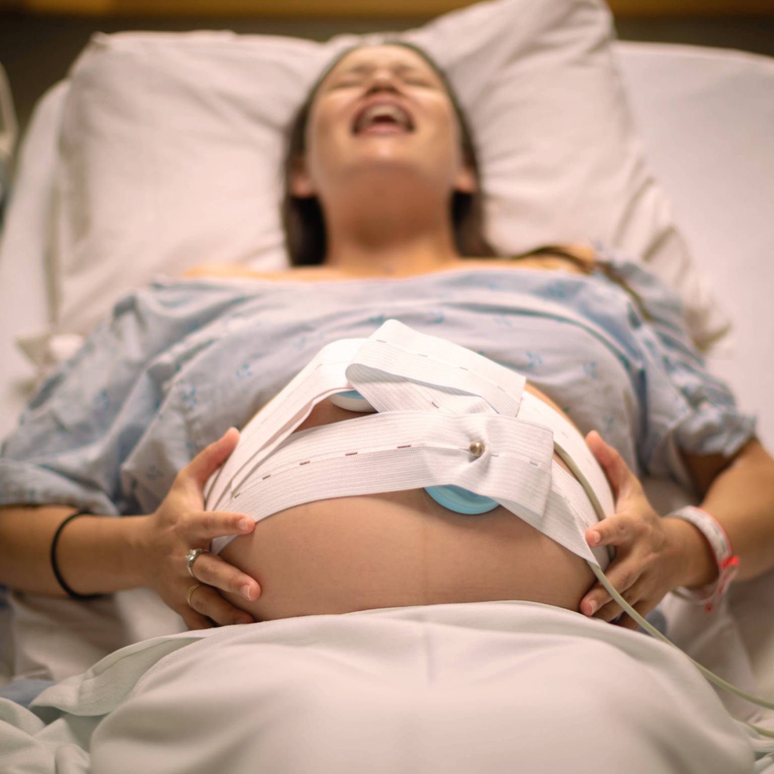 Schwangere Frau im Krankenhausbett, trägt ein Krankenhaushemd, hat Wehen und hat ein Gerät zur fetalen Überwachung auf ihrem Bauch.