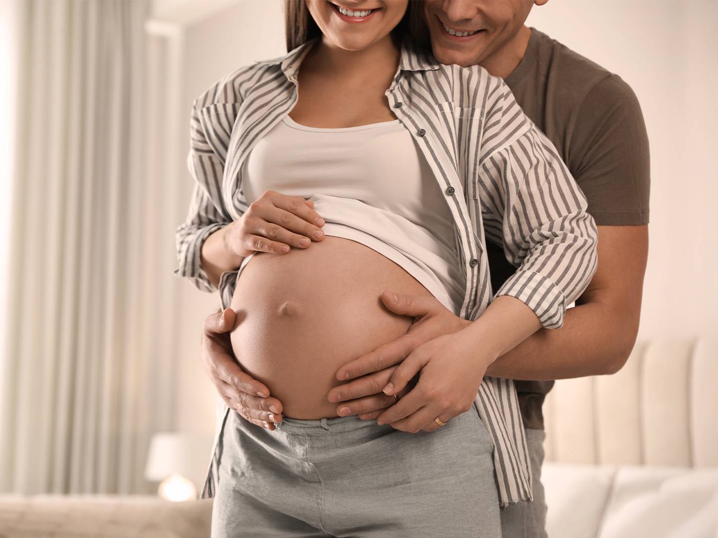 Eine schwangere Frau und ein Mann stehen lächelnd nebeneinander. Der Mann hält sanft ihren Bauch und symbolisiert Liebe und Vorfreude in einem gemütlichen Raum.