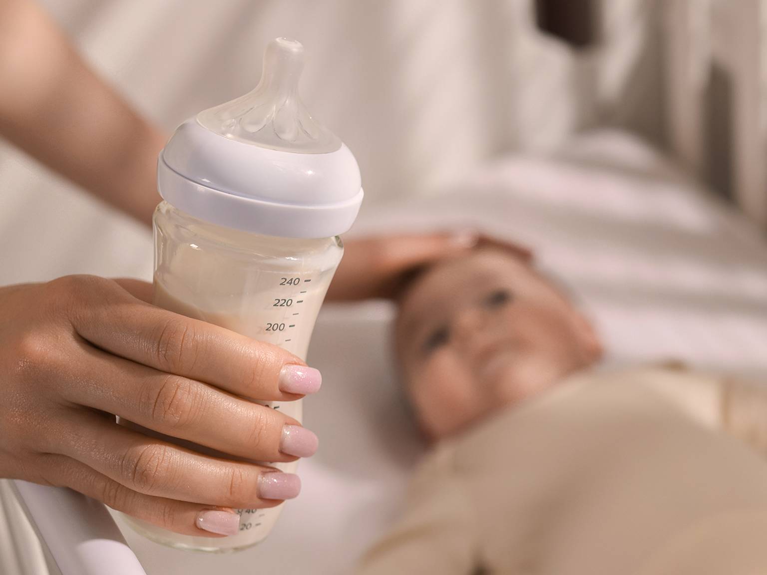 Eine Hand hält eine mit Milch gefüllte Babyflasche neben einem Baby, das in einem Kinderbett liegt, und berührt sanft den Kopf des Babys.