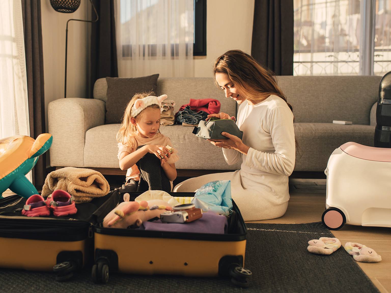 Eine Frau und ein Kind packen einen Koffer in einem gemütlichen Wohnzimmer, umgeben von Kleidung und Spielzeug, um sich auf eine Reise vorzubereiten.