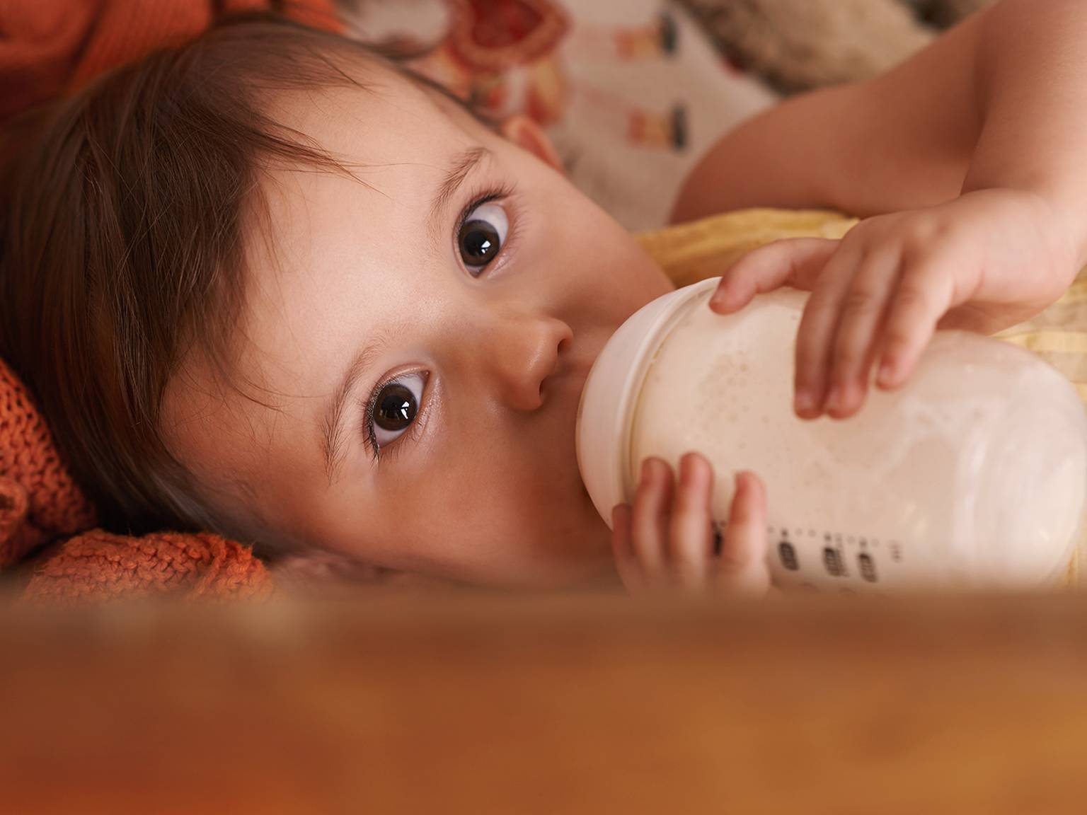 Baby liegt, trinkt aus der Flasche, hat große Augen und eine kuschelige Decke.
