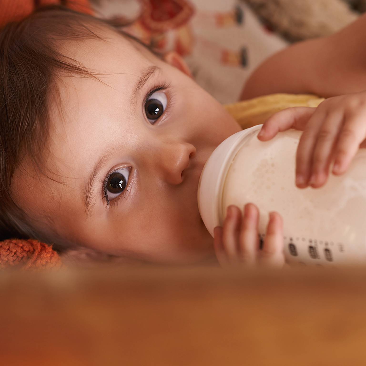 Baby liegt, trinkt aus der Flasche, hat große Augen und eine kuschelige Decke.