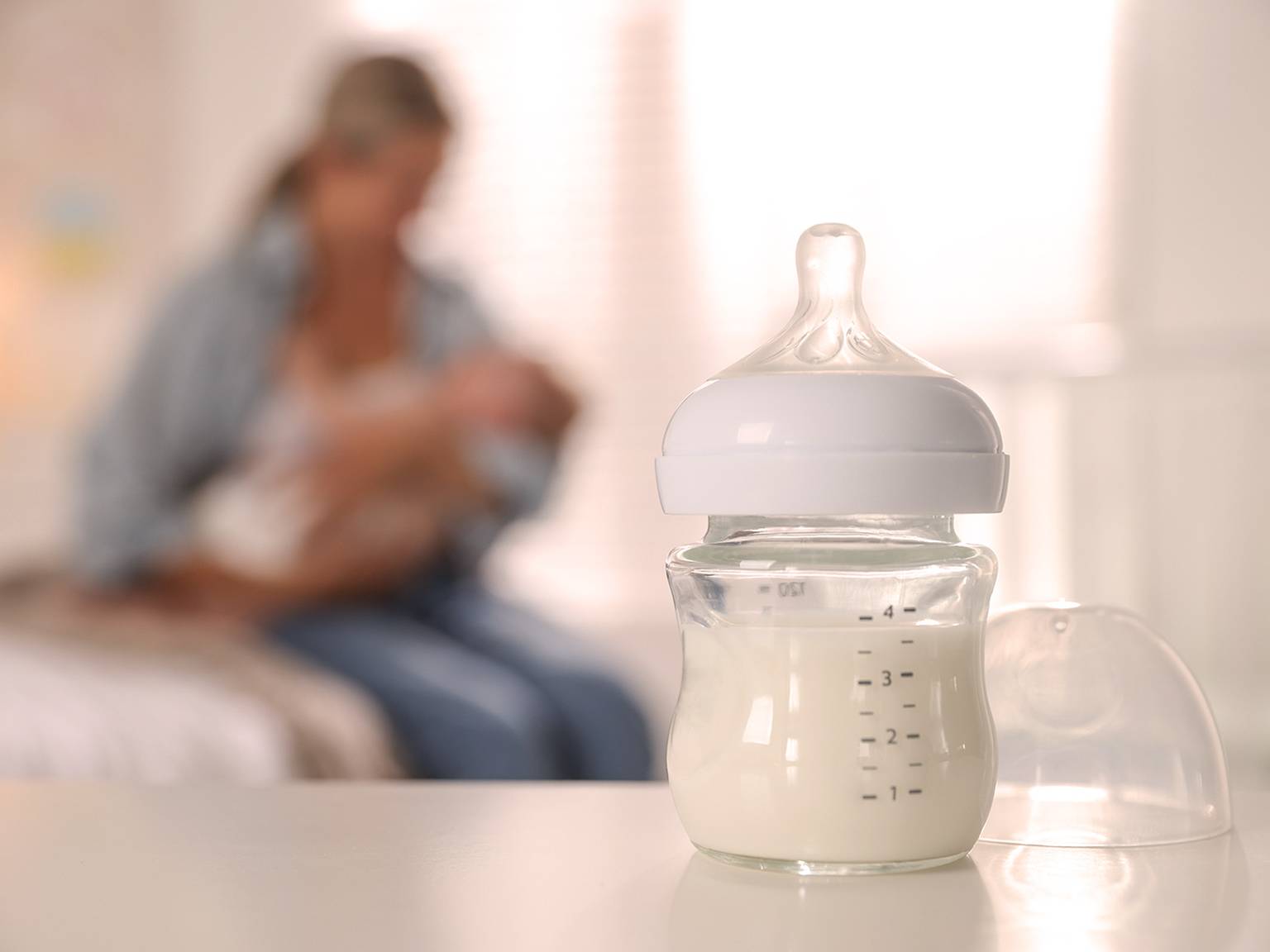 Auf einem Tisch steht eine mit Milch gefüllte Babyflasche. Im unscharfen Hintergrund hält eine Person ein Baby.