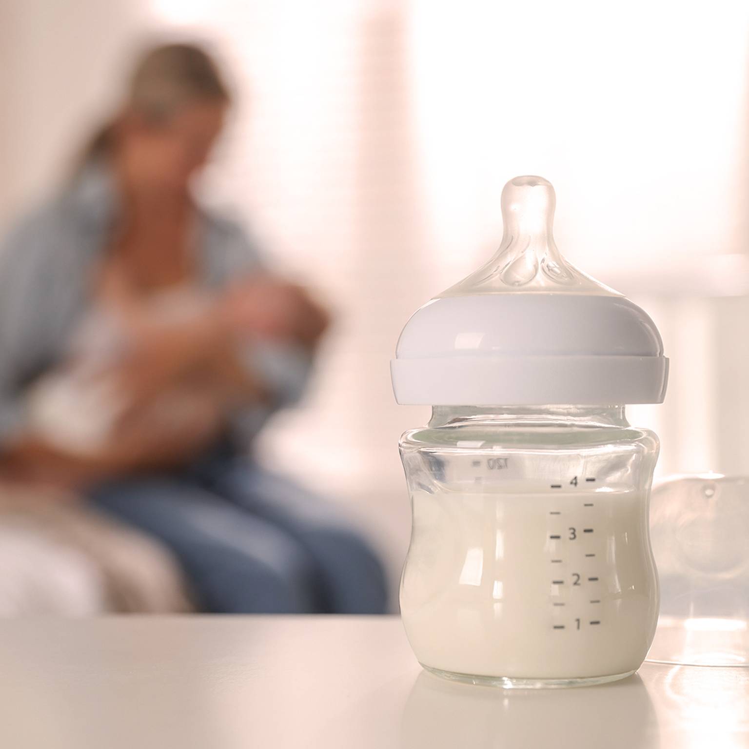 Auf einem Tisch steht eine mit Milch gefüllte Babyflasche. Im unscharfen Hintergrund hält eine Person ein Baby.